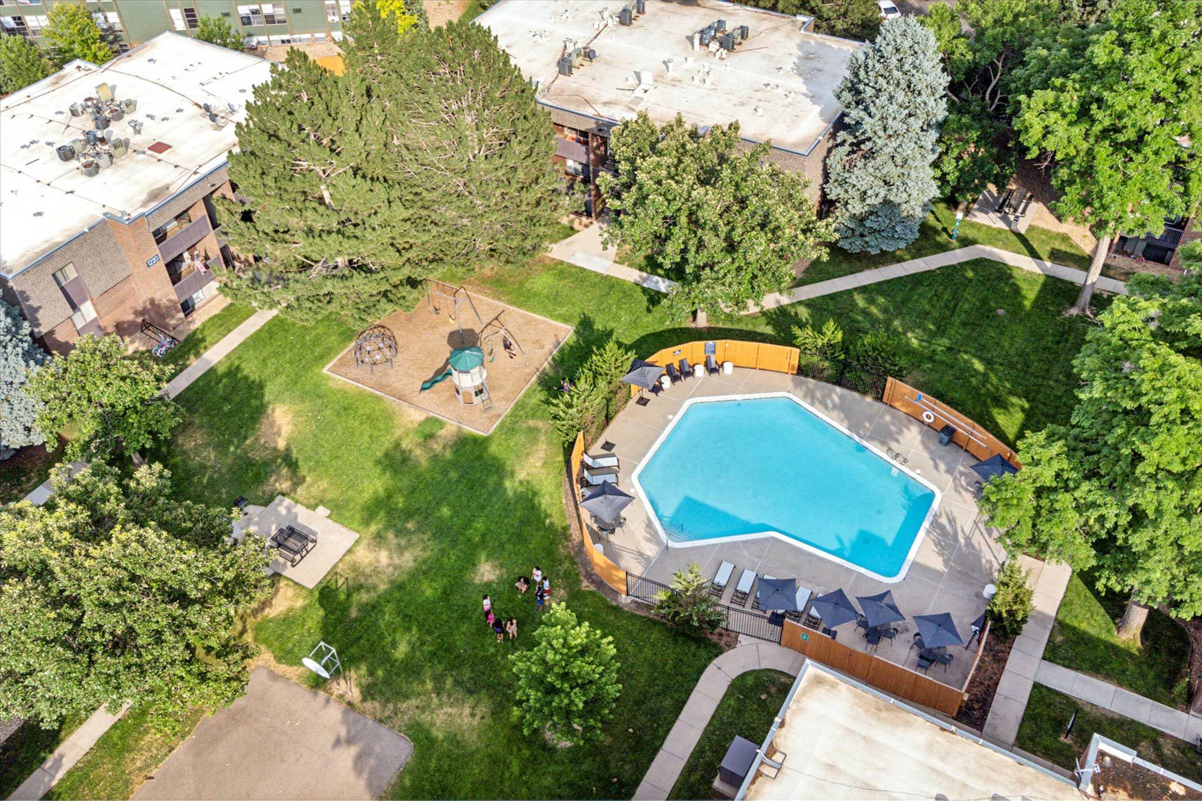 An aerial view of a residential complex featuring a blue swimming pool, a playground with a swing set, and well-maintained green lawns surrounded by trees. Several lounge chairs are arranged around the pool, and a small group of people can be seen nearby. The buildings have flat roofs and are set within a landscaped area.