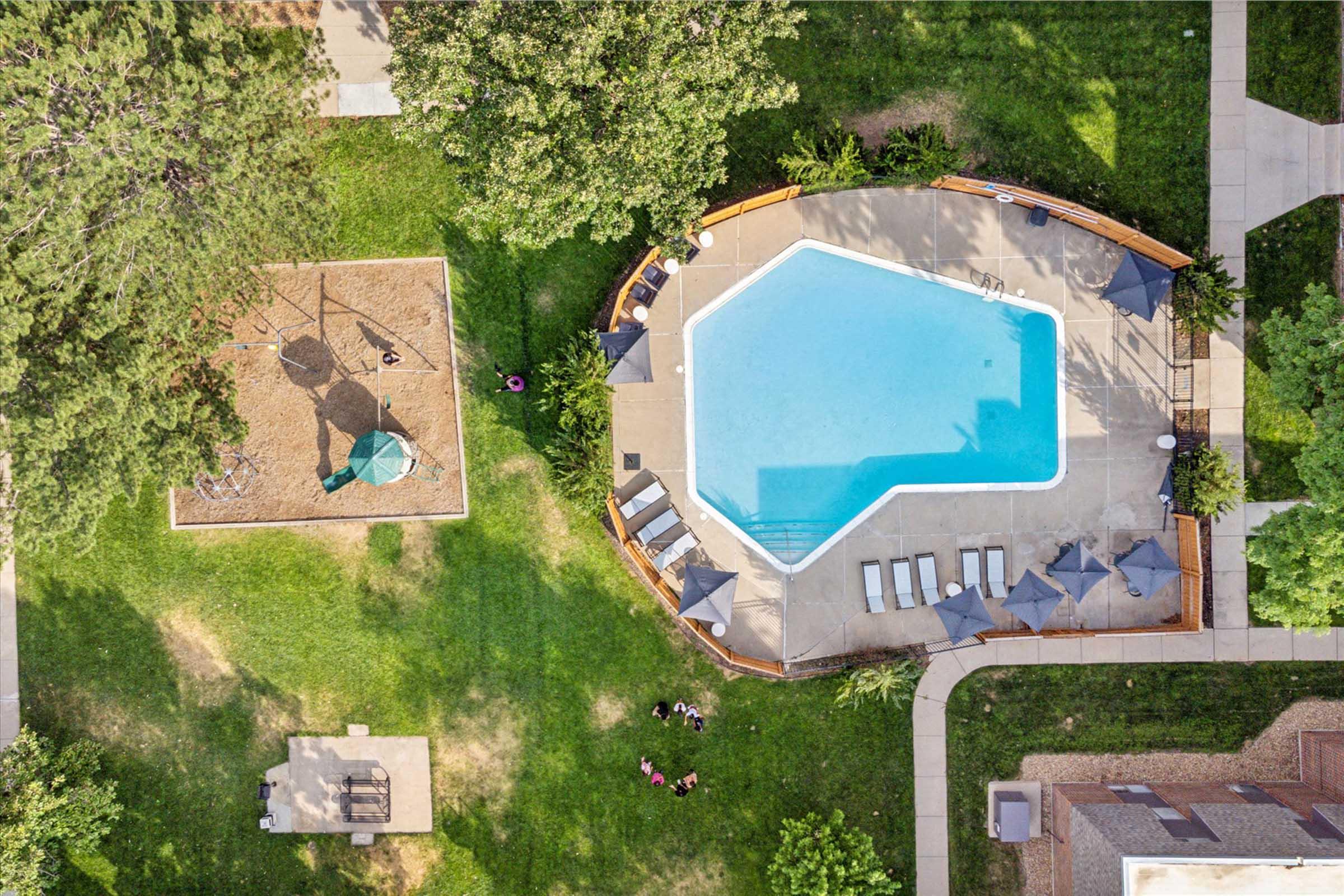 Aerial view of a swimming pool with an octagonal shape surrounded by sun loungers and umbrellas. Nearby, there is a playground with swings and a slide on the left, and a grassy area with a few people relaxing on the right. Trees line the perimeter, enhancing the outdoor atmosphere.