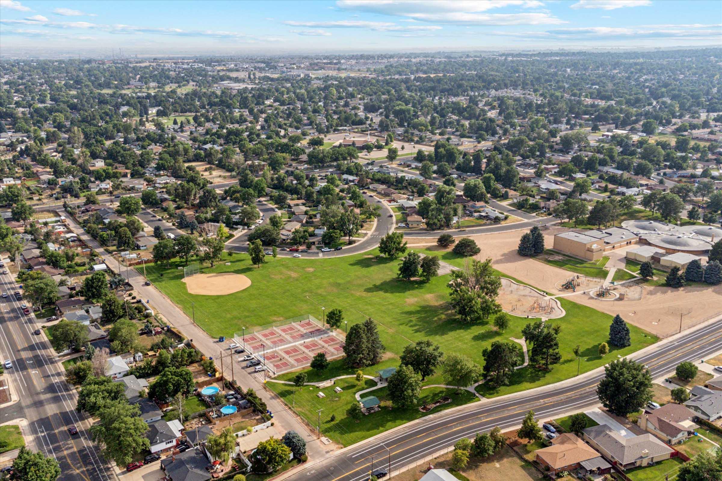 Aerial view of a suburban neighborhood featuring a large green park with baseball and basketball facilities, surrounded by residential homes. The park has trees, open grassy areas, and playground equipment visible. Streets run alongside the neighborhood, showcasing a mix of houses and local amenities.