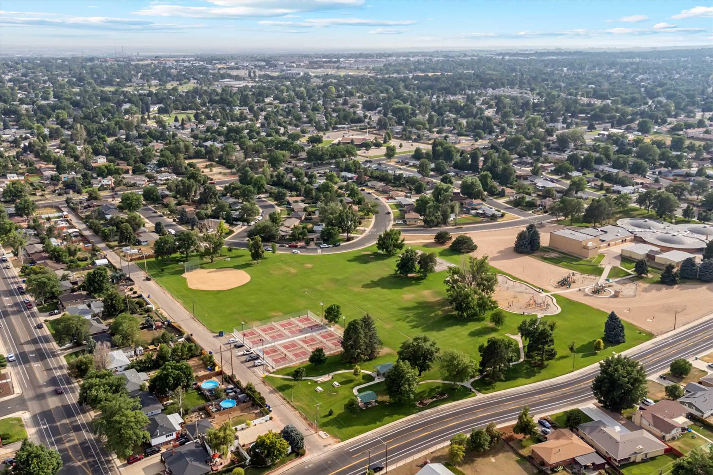 Aerial view of a residential area featuring a large green park with baseball and basketball courts, playgrounds, and surrounding trees. Nearby streets are lined with houses, and the skyline is visible in the background under a clear blue sky.