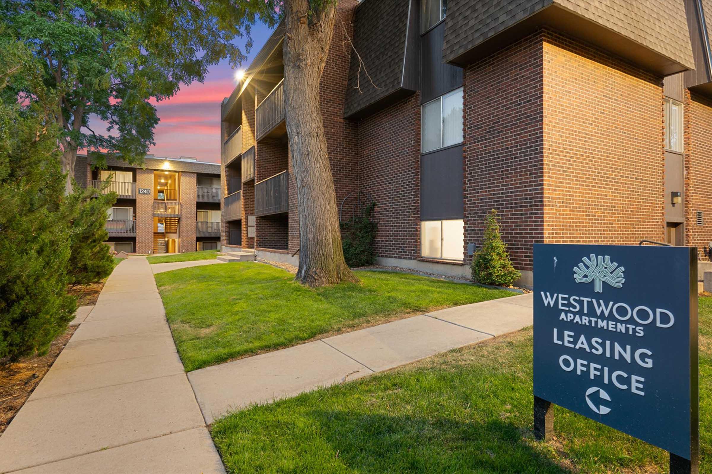 A pathway leads to the Westwood Apartments leasing office, flanked by green lawns and trees. The building features a mix of brick and modern architecture, with an illuminated entrance in the background. A sign for the leasing office is prominently displayed. The sky shows a colorful sunset.