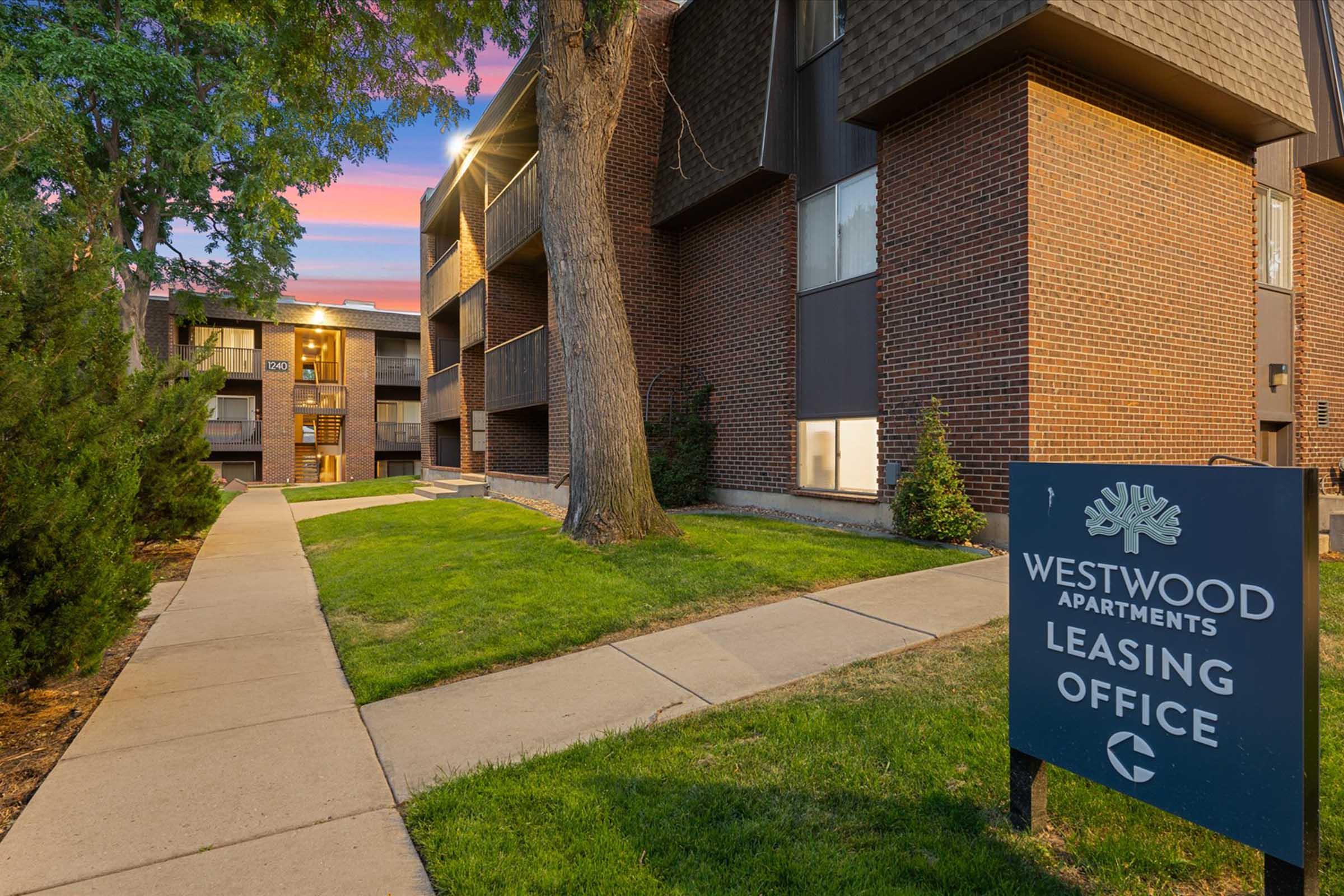 A pathway leading to the Westwood Apartments leasing office, featuring modern brick architecture and large windows. Lush green lawns flank the walkway, and a prominent sign displaying "Westwood Apartments Leasing Office" is visible in the foreground. The sky shows a pastel sunset.
