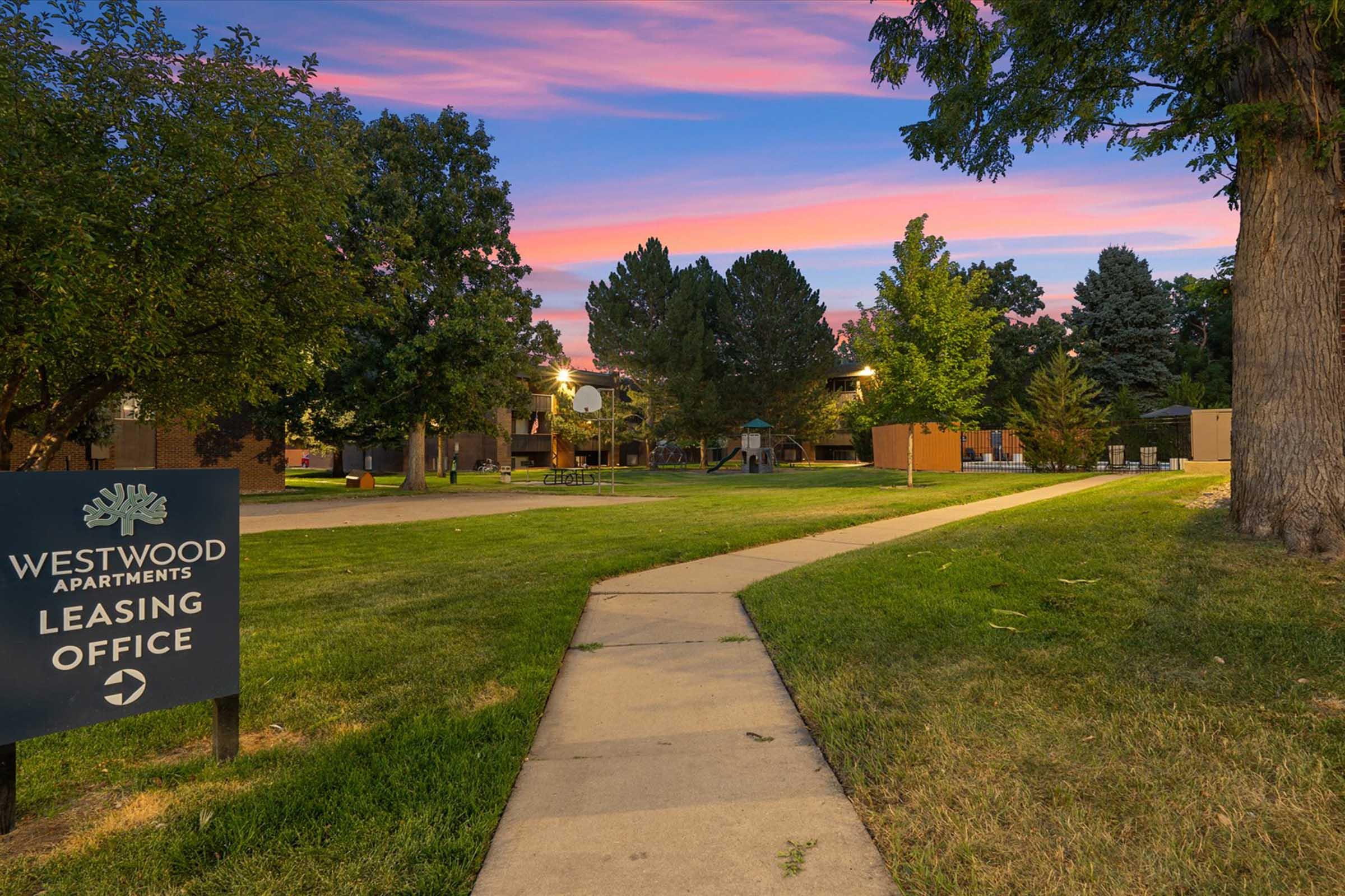 Pathway leading to the leasing office of Westwood Apartments, surrounded by trees and a grassy area. The sky is painted with pink and blue hues at sunset, creating a serene atmosphere. A sign indicates the direction to the leasing office.