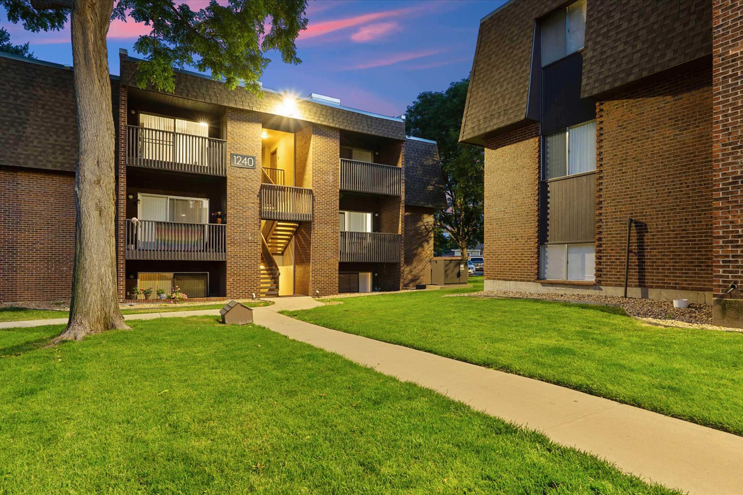 A view of a multi-unit residential building at dusk, featuring brick exteriors with balconies. A well-maintained lawn and pathway are visible in the foreground, along with trees and ambient lighting that highlights the architecture and landscaping. The scene conveys a welcoming, peaceful atmosphere.