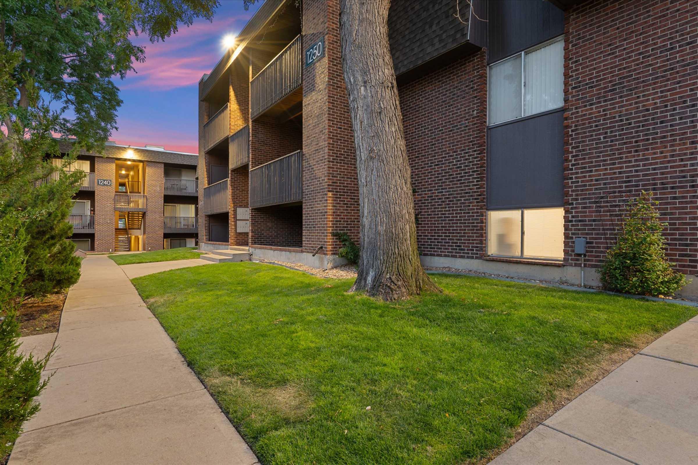 Exterior view of an apartment complex featuring brick buildings, a paved walkway, and landscaped green areas. The scene includes trees and shrubs, with some outdoor lighting visible, along with a colorful sky at dusk.