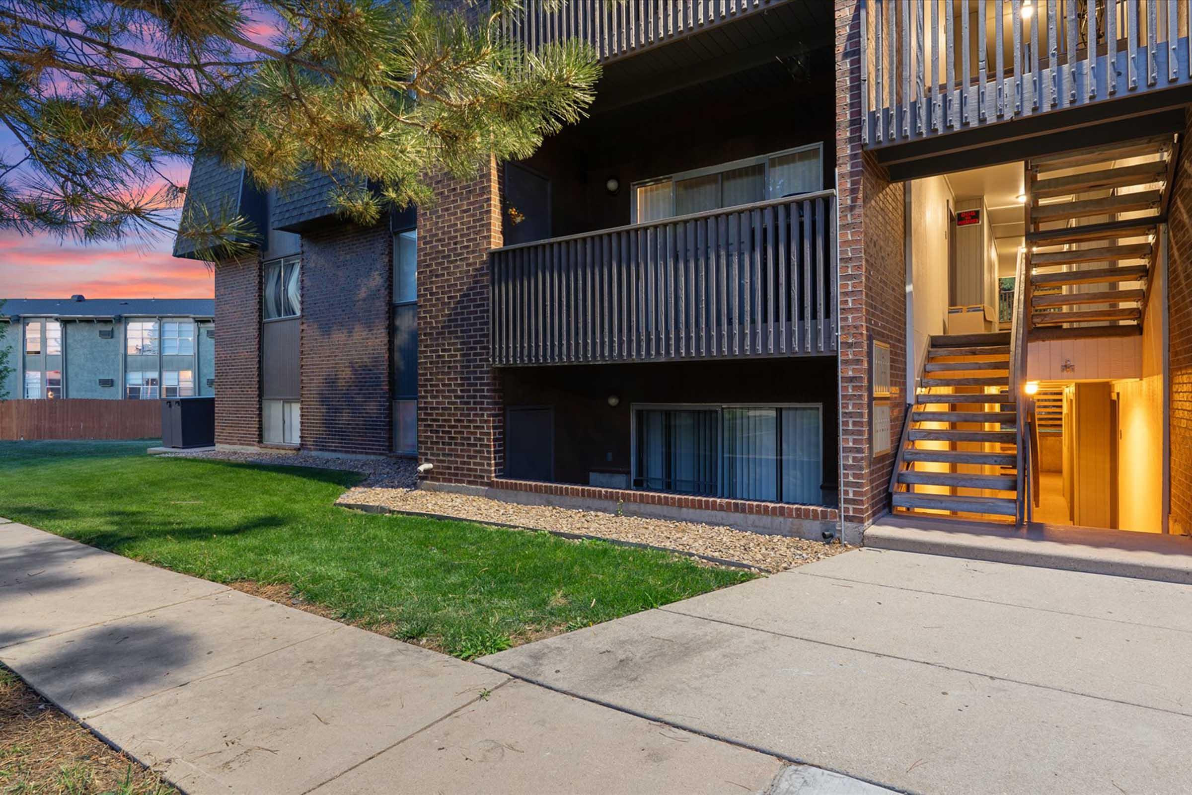 Exterior view of an apartment building at sunset, featuring a brick facade and wooden balcony railings. A staircase leads to an upper floor, with a well-maintained lawn in the foreground. The overall atmosphere is inviting and well-kept, showcasing accessible entrance points.