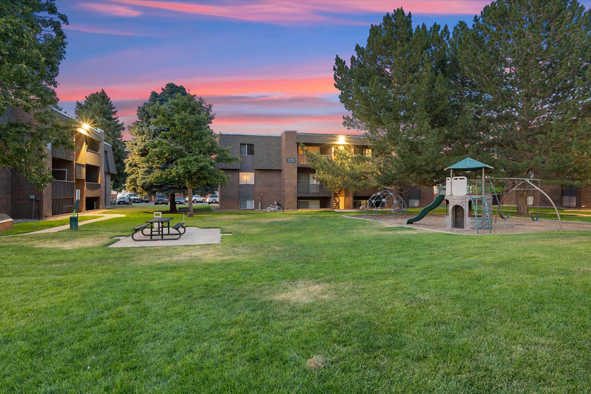 A landscaped courtyard of an apartment complex at sunset, featuring green grass, trees, and a playground structure. Picnic tables are visible in the foreground, with the building and parking area in the background, under a colorful sky.