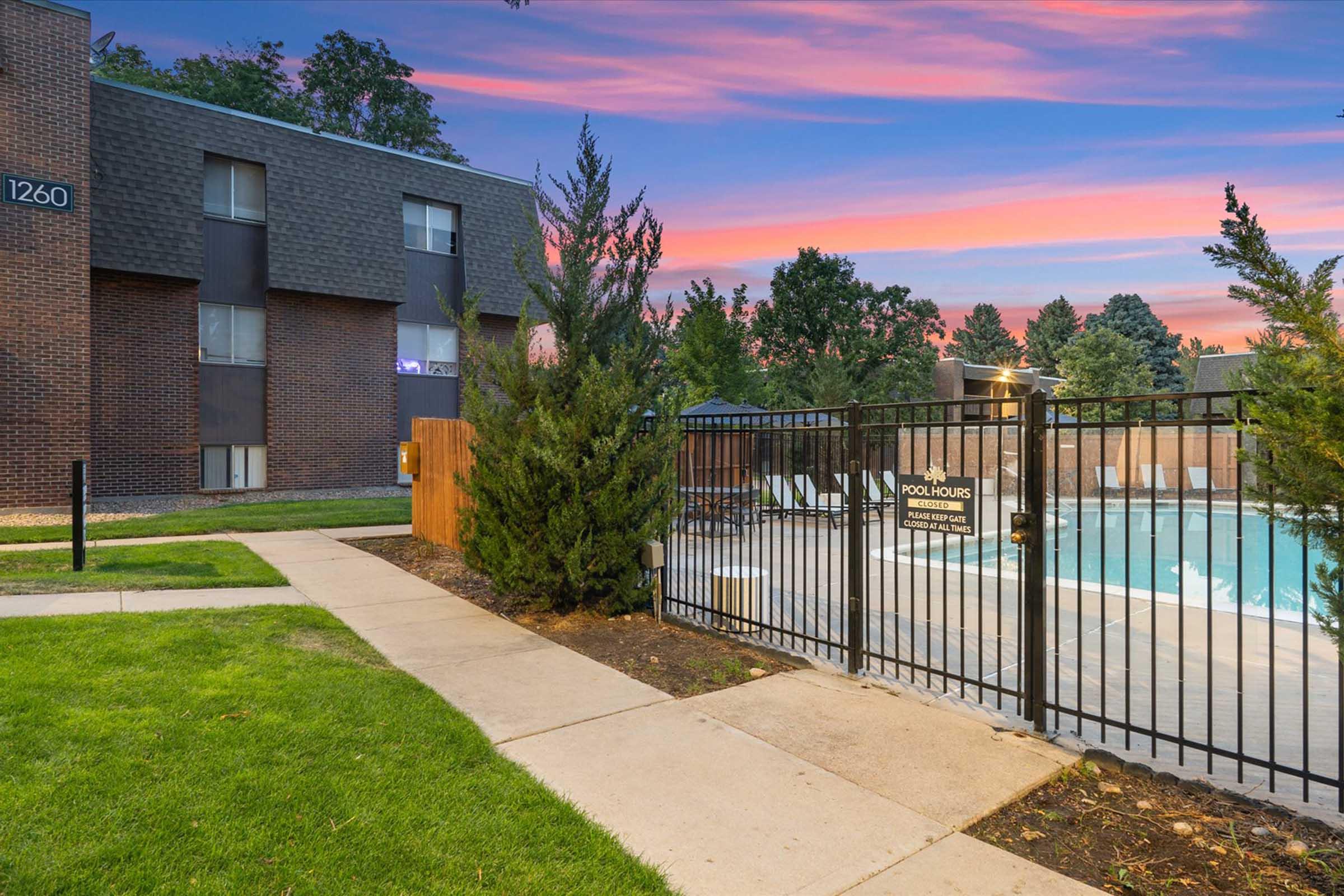 A serene pool area at sunset, featuring a fenced pool with chairs and umbrellas. Lush green grass lines the path leading to the pool, while trees provide a natural border. The sky showcases vibrant hues of pink and purple, enhancing the tranquil ambiance of the scene.