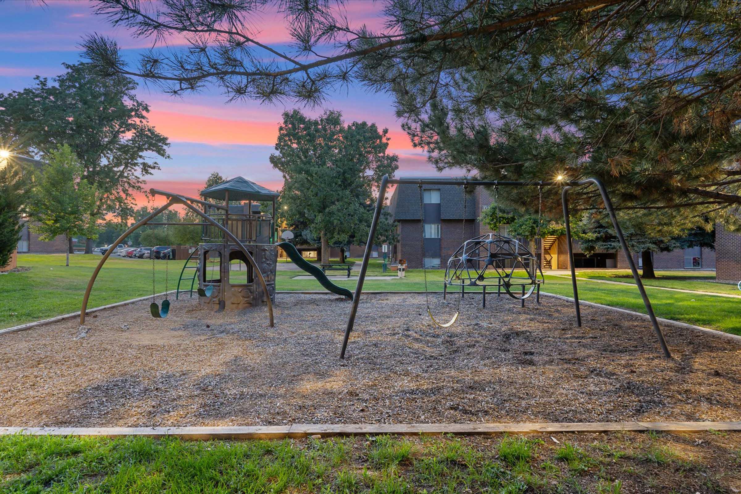 A playground area featuring swings, a climbing structure, and a slide, set in a grassy space. Surrounding trees provide shade, and residential buildings are visible in the background under a colorful sunset sky.