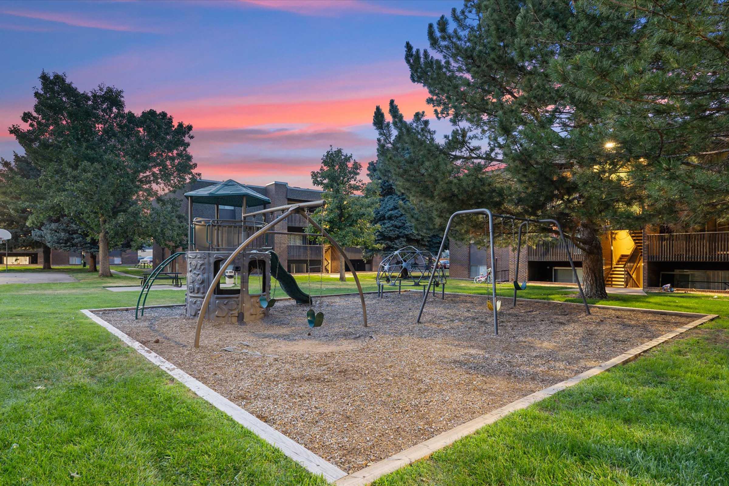 Playground scene at sunset featuring swings, climbing structures, and slides surrounded by trees and grass. Residential buildings are visible in the background under a colorful sky. The area is well-maintained, providing a playful atmosphere for children.