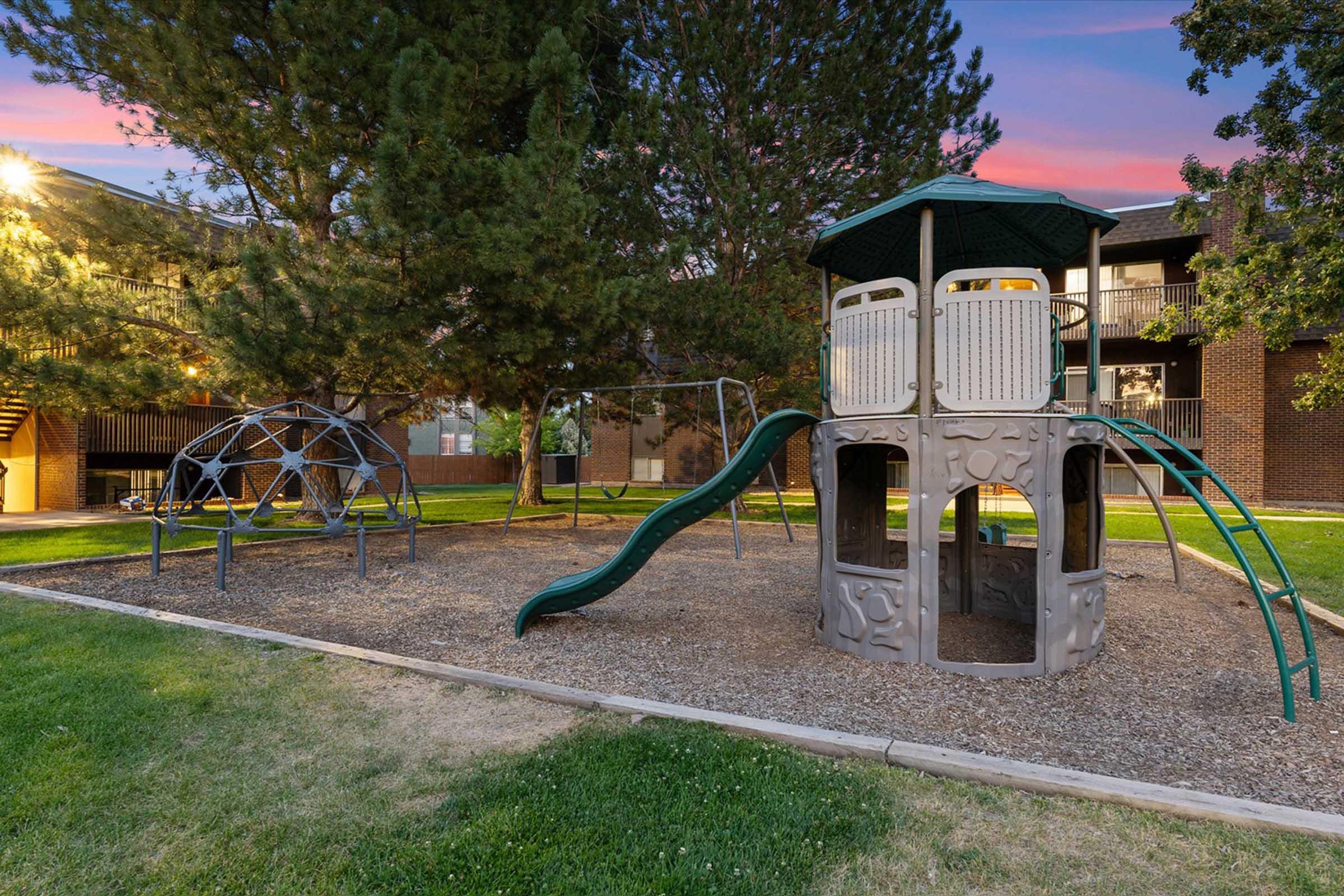 A playground featuring a climbing structure with a slide, surrounded by grass and trees. In the background, there are outdoor apartments illuminated by warm lighting. The scene depicts a recreational area for children, offering safe play opportunities in a community setting.
