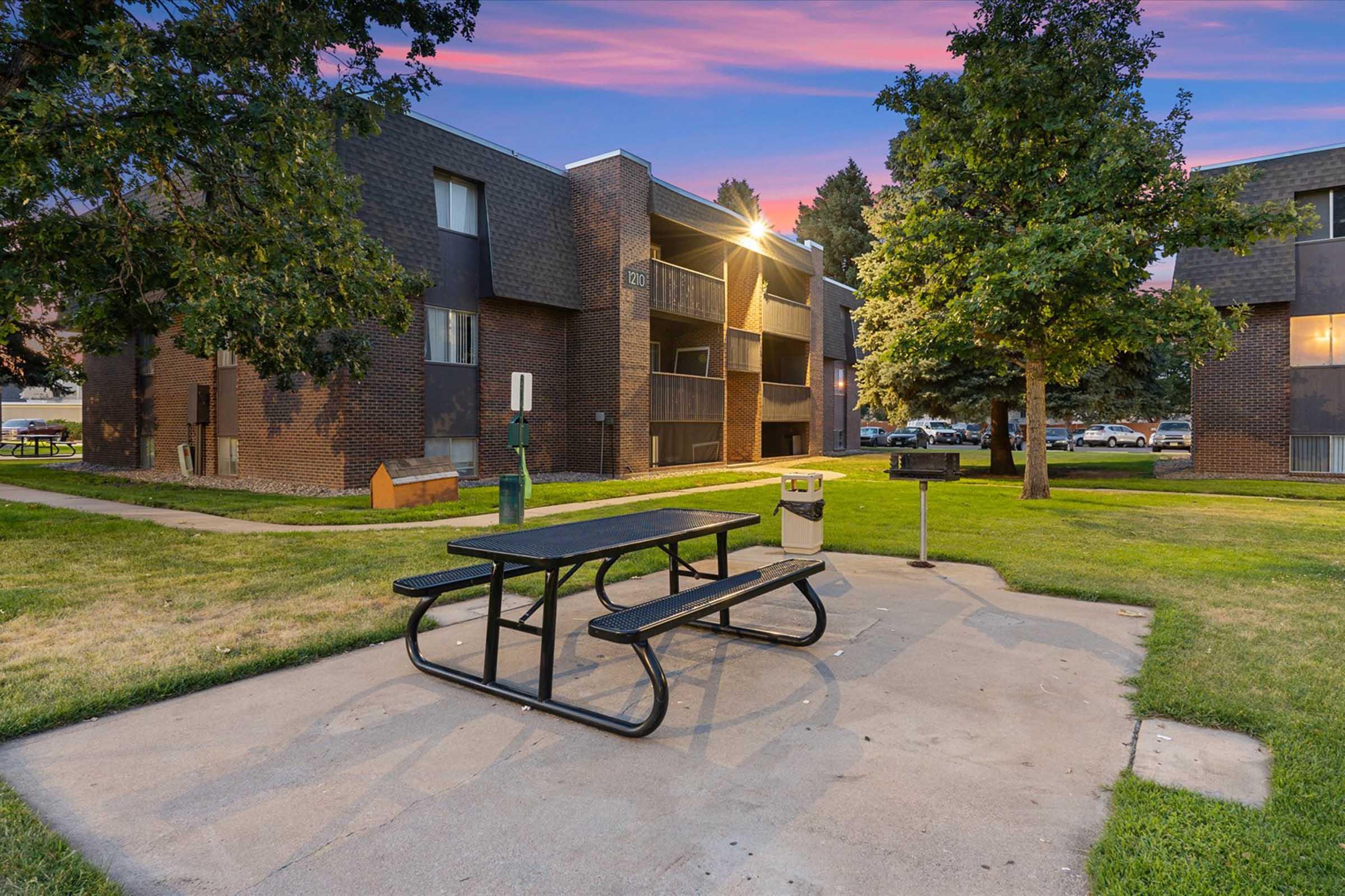 A picnic area featuring a black metal table and benches on a concrete slab, surrounded by grassy lawns and trees. In the background, a two-story brick apartment building is visible, with lights on in some of the units. The sky is painted with soft pink and purple hues, indicating dusk.