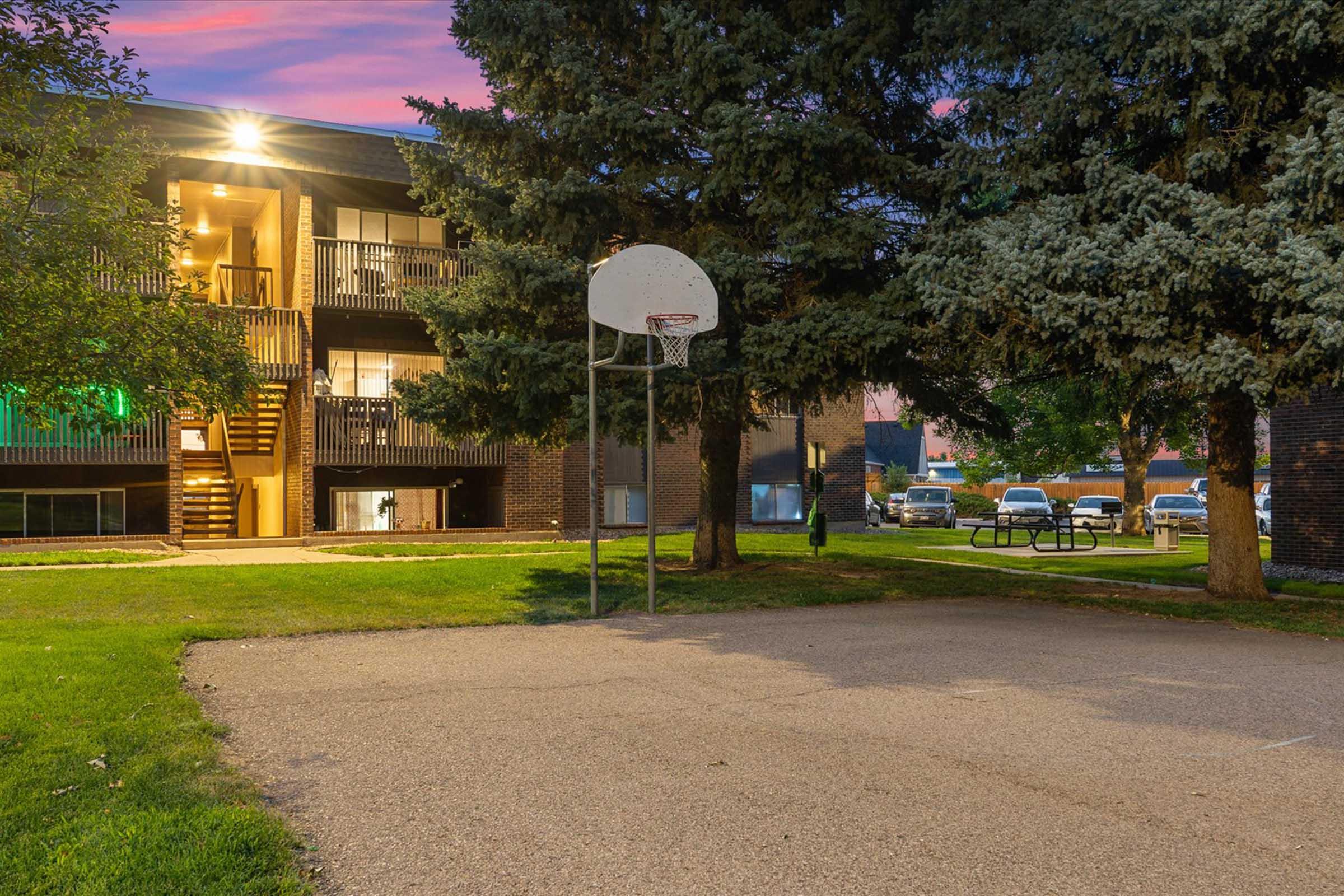 A basketball hoop stands on a paved court surrounded by grassy areas and large trees. In the background, an apartment building with illuminated balconies can be seen, along with parked cars and a picnic area. The sky is painted in hues of purple as the day transitions to evening.