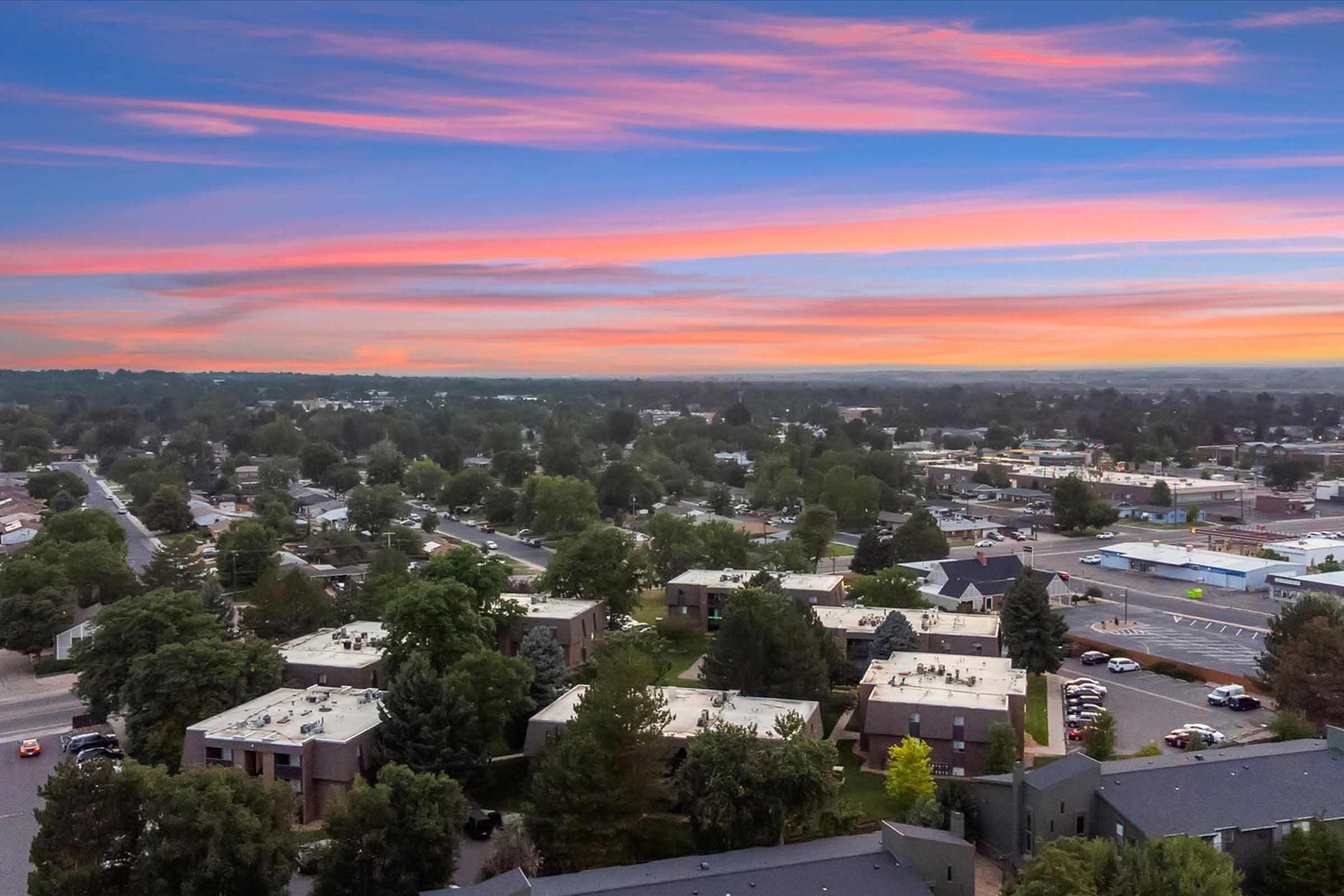 Aerial view of a suburban area featuring residential buildings and tree-lined streets. The sky is painted with soft pink and blue hues during sunset, creating a tranquil atmosphere. In the distance, a commercial area is visible, blending with the greenery of the landscape.