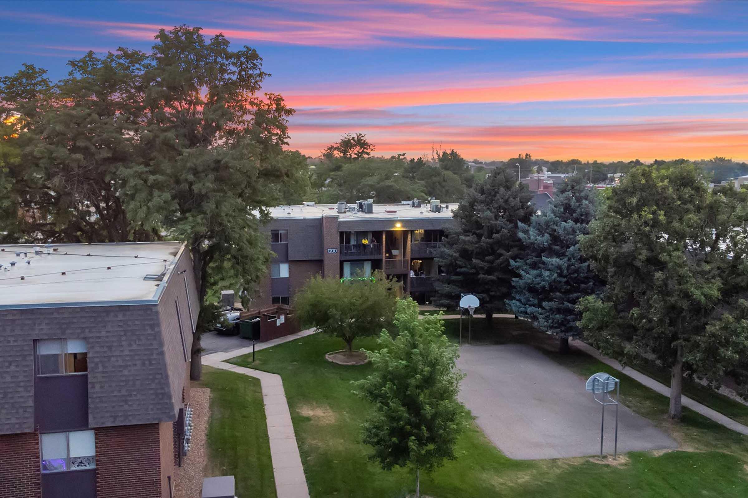 A residential area featuring multi-story apartment buildings and a basketball hoop. Lush trees are scattered throughout the property, and the sky displays a vibrant sunset with shades of pink and purple. The scene conveys a peaceful suburban atmosphere.