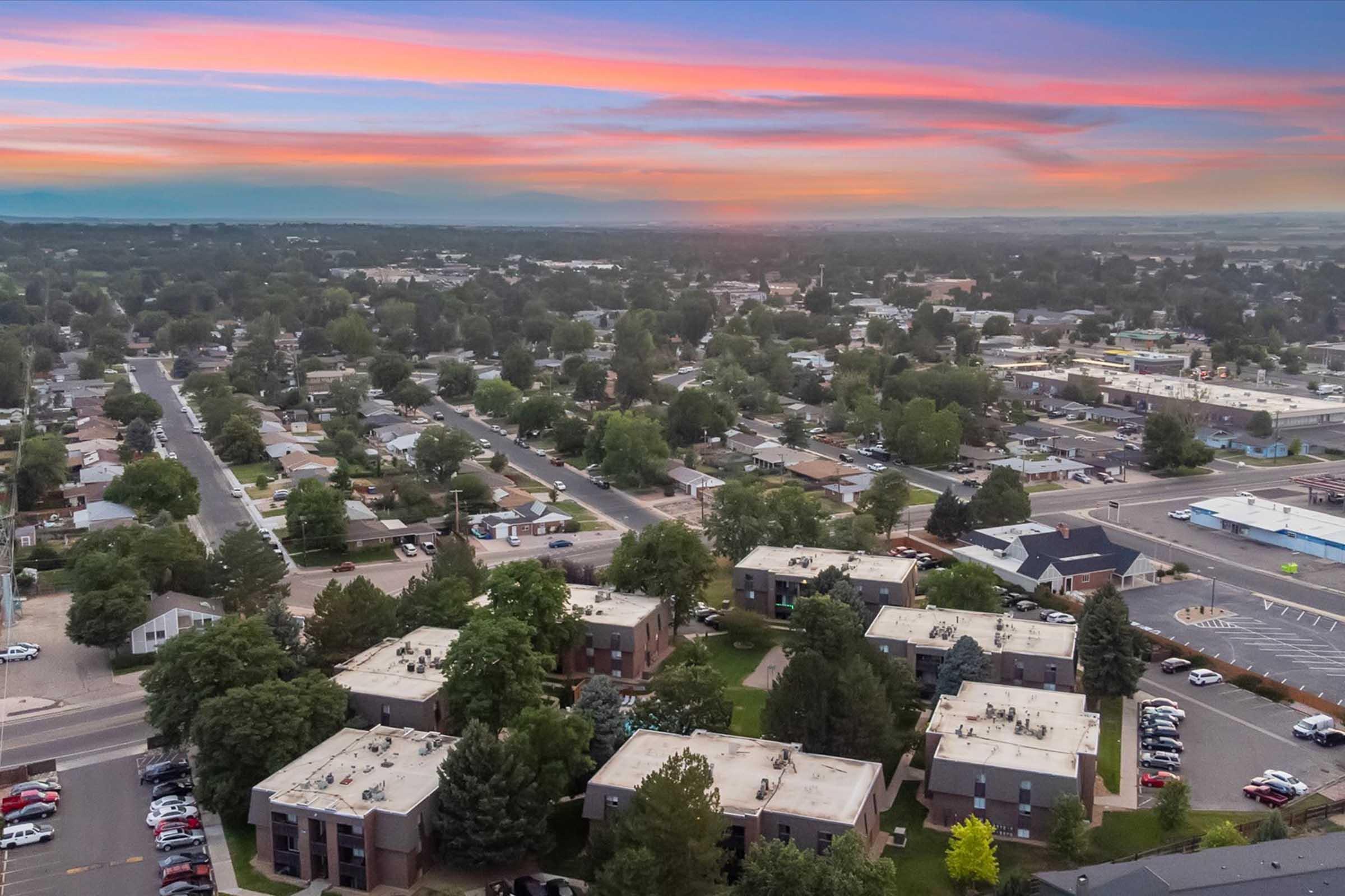 Aerial view of a suburban neighborhood during sunset. The image features a mix of residential buildings, trees, and streets, with a colorful sky displaying shades of pink, orange, and blue. In the foreground, several apartment complexes and parking lots are visible, surrounded by greenery and a backdrop of distant hills.