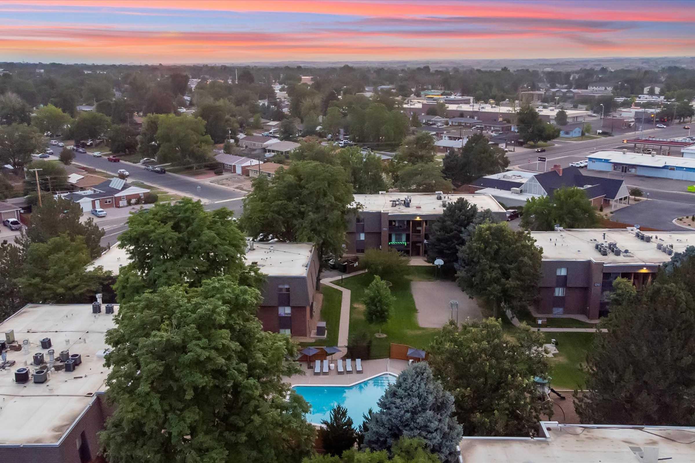 Aerial view of a residential area featuring several apartment buildings surrounded by trees and a swimming pool. The sky displays a colorful sunset with shades of orange and pink, while the neighborhood includes streets and smaller buildings in the background.
