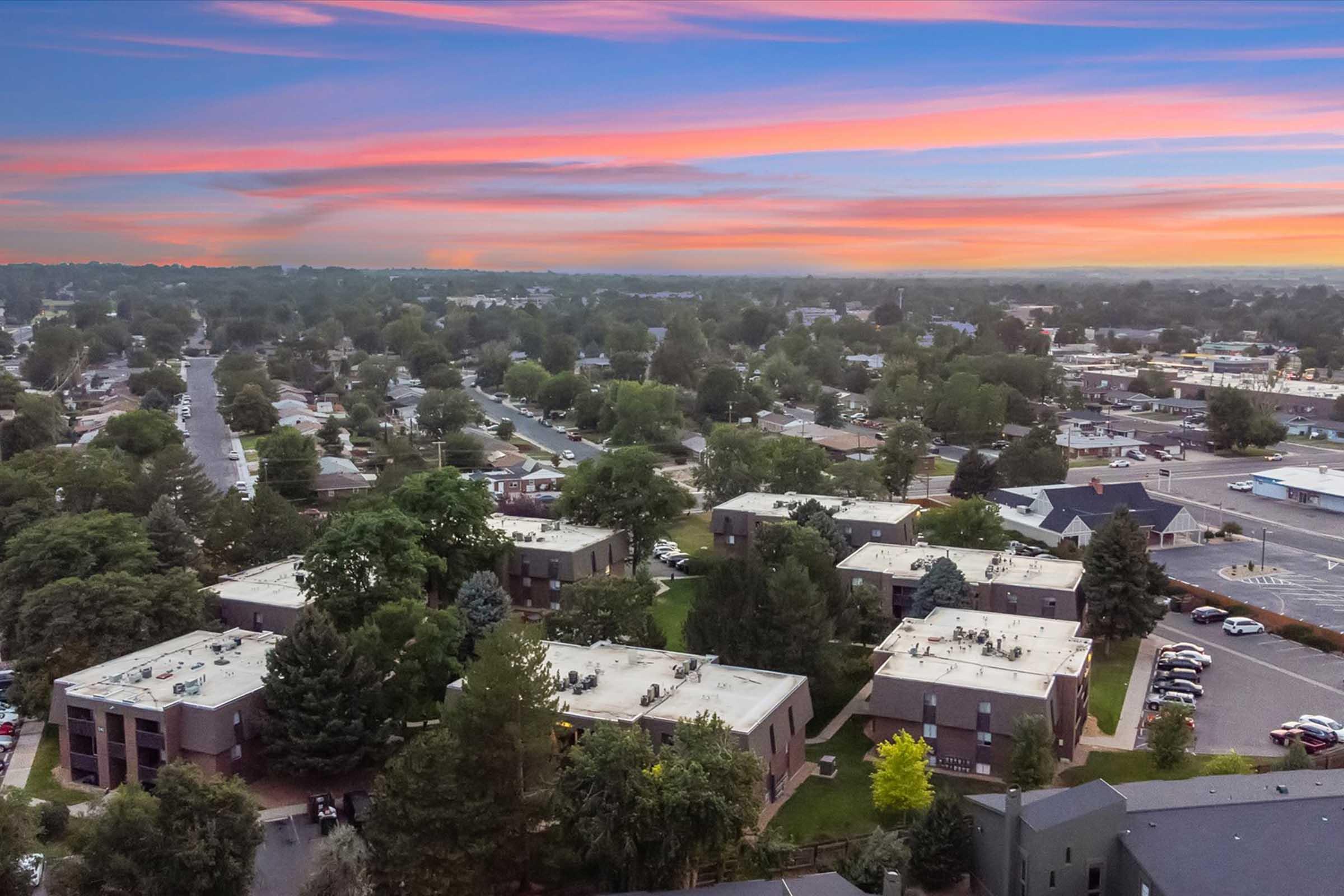 Aerial view of a suburban neighborhood at sunset, featuring several residential buildings surrounded by trees, green spaces, and roads. The sky displays vibrant colors with streaks of pink, orange, and purple, enhancing the tranquil atmosphere of the community below.