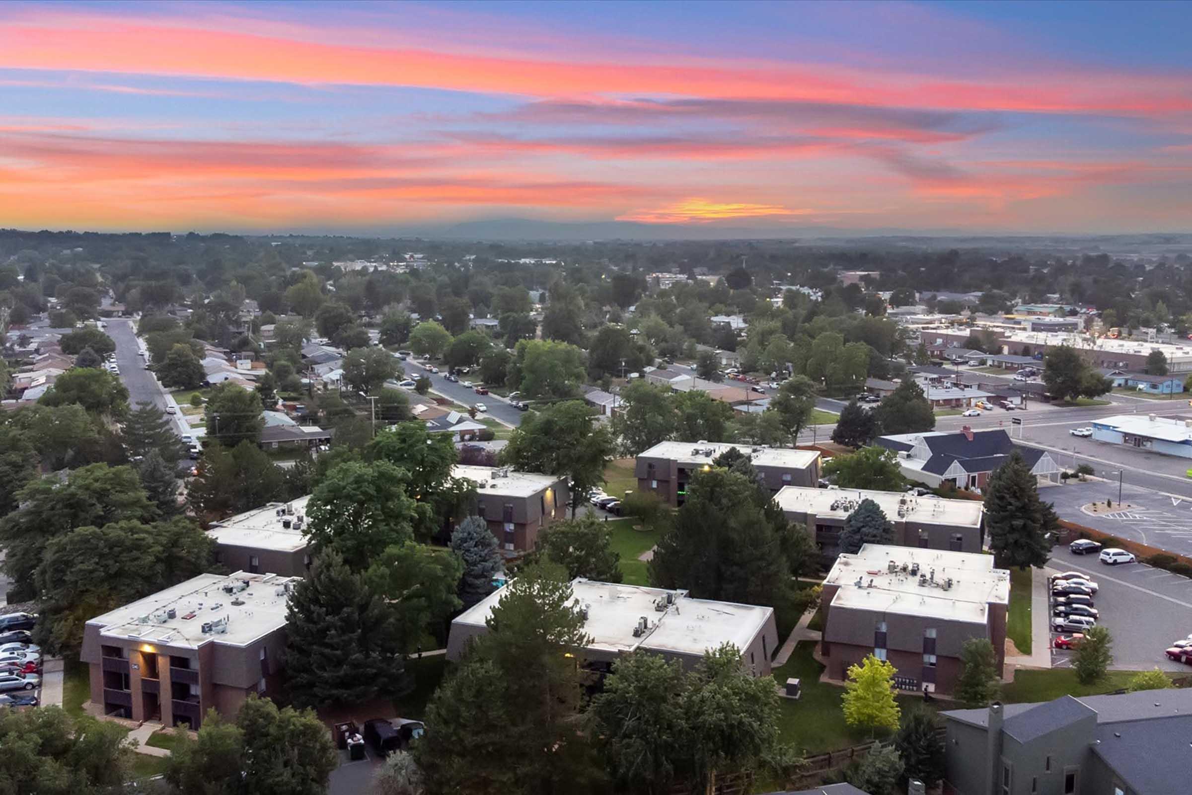 Aerial view of a suburban neighborhood at sunset, featuring clusters of apartment buildings surrounded by trees, with a colorful sky. The layout shows roads and nearby shops, depicting a peaceful residential area.