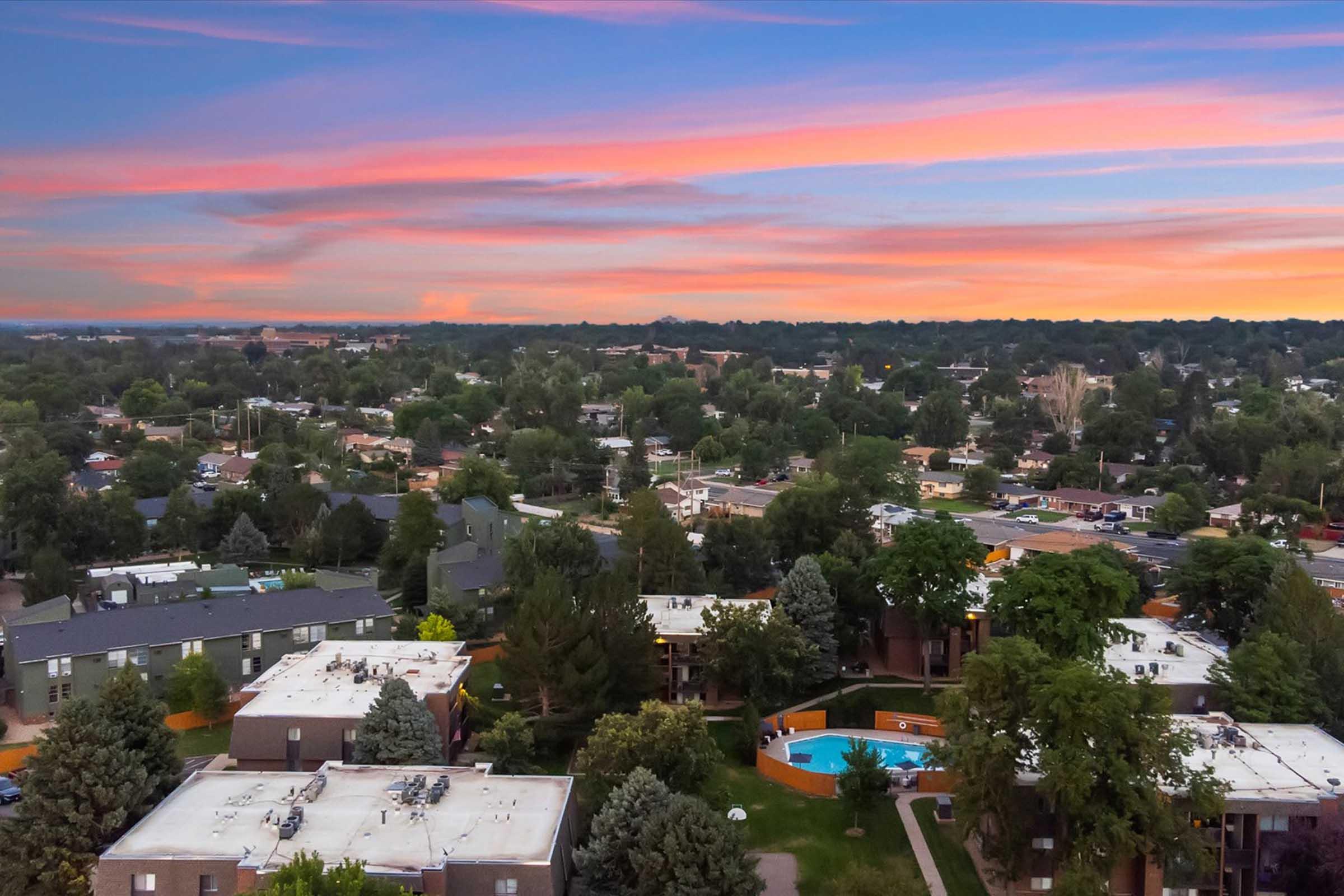 Aerial view of a suburban neighborhood at sunset, featuring residential buildings surrounded by trees. The sky displays vibrant pink and orange hues, complementing the green landscape and pools in the area. The scene conveys a peaceful, picturesque atmosphere.