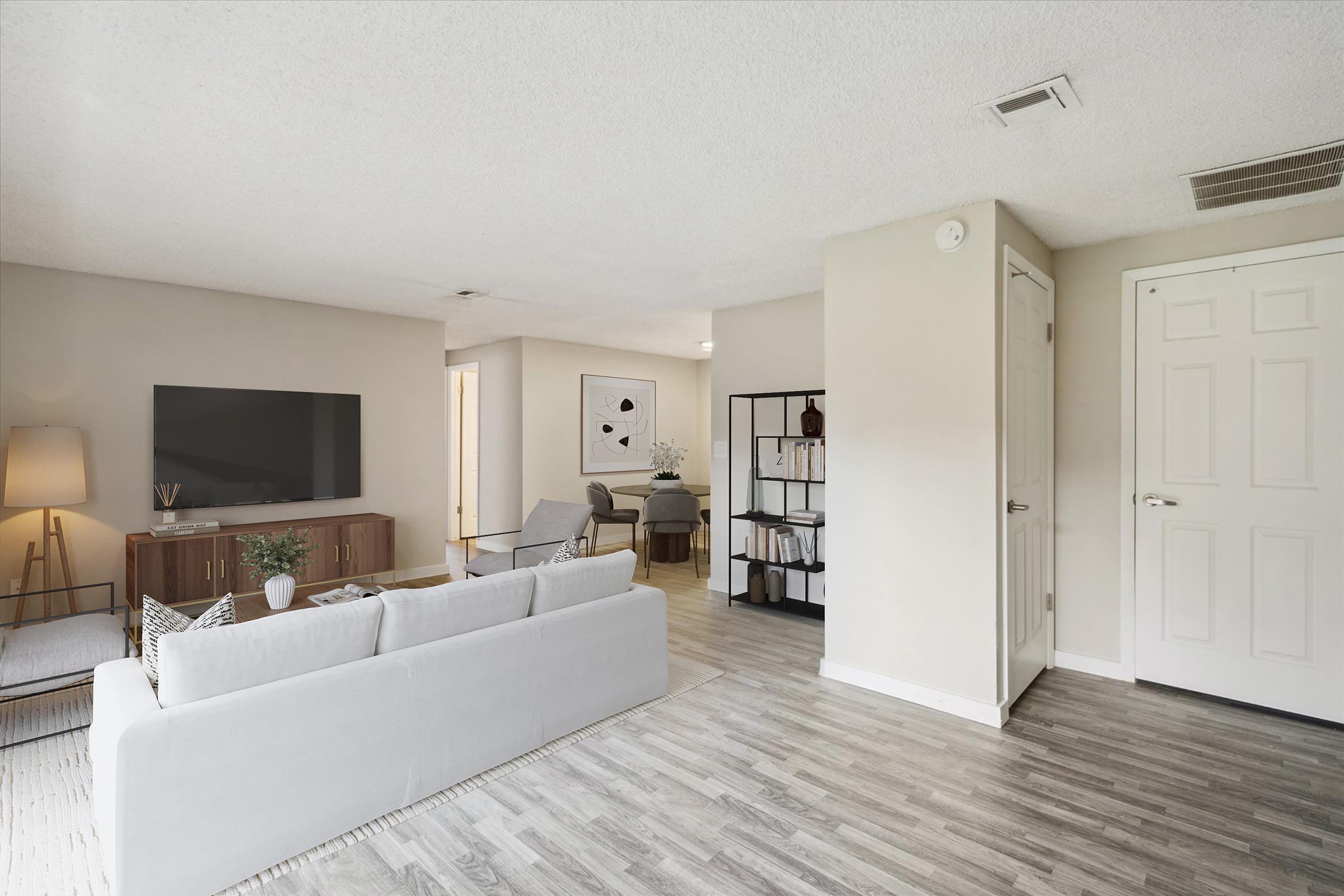 A modern living room featuring a light-colored sofa, a wooden TV stand with a flat-screen TV, and a stylish lamp. The space is decorated with a plant and has neutral walls. In the background, a cozy reading chair and a bookshelf can be seen, along with an open doorway leading to another room.