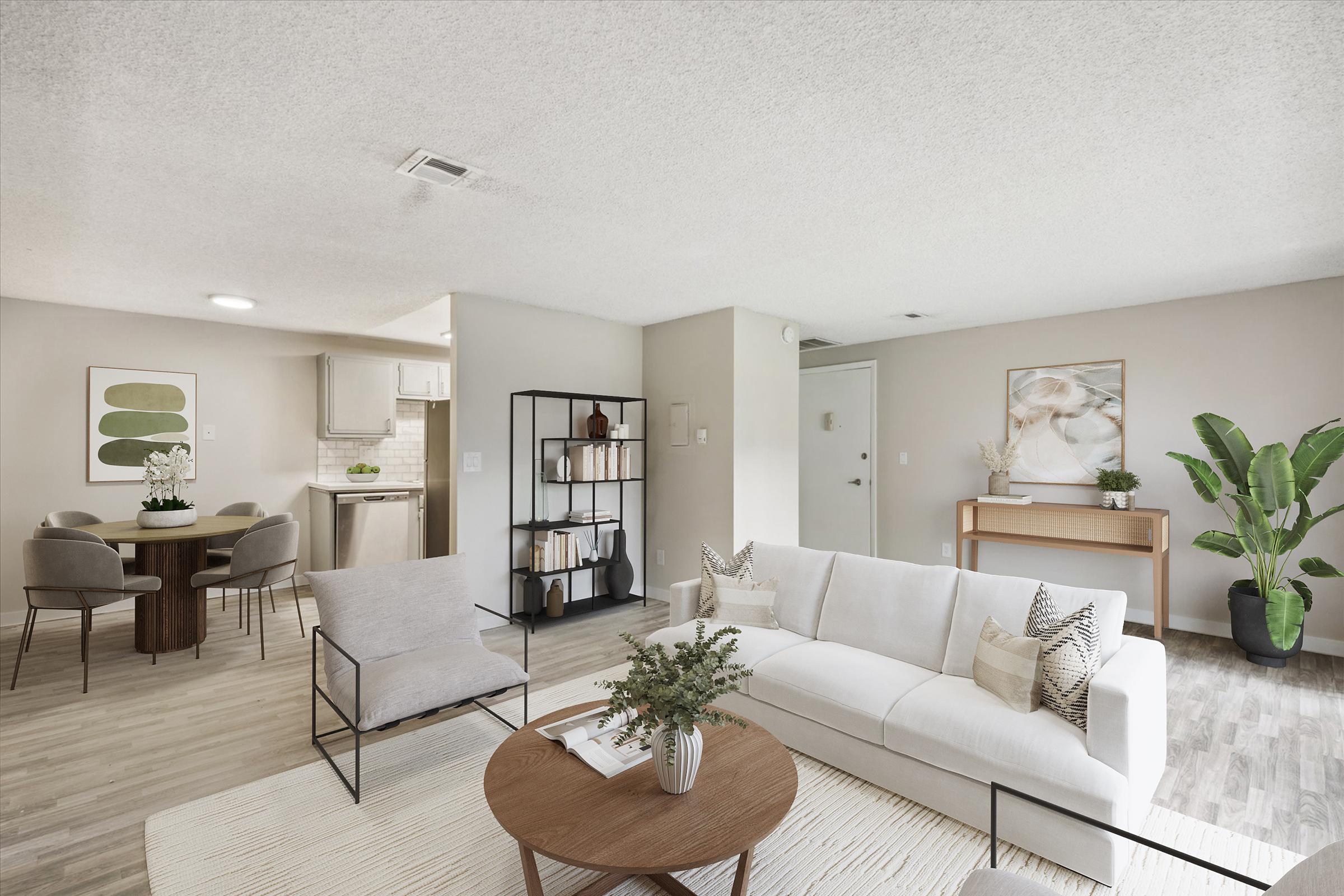 A bright, modern living room featuring a white sofa, a round coffee table with a small plant, and two armchairs. There’s a dining area in the background with a table and chairs. Decorative plants and artwork add warmth to the space. The flooring is light-colored wood, creating an inviting atmosphere.