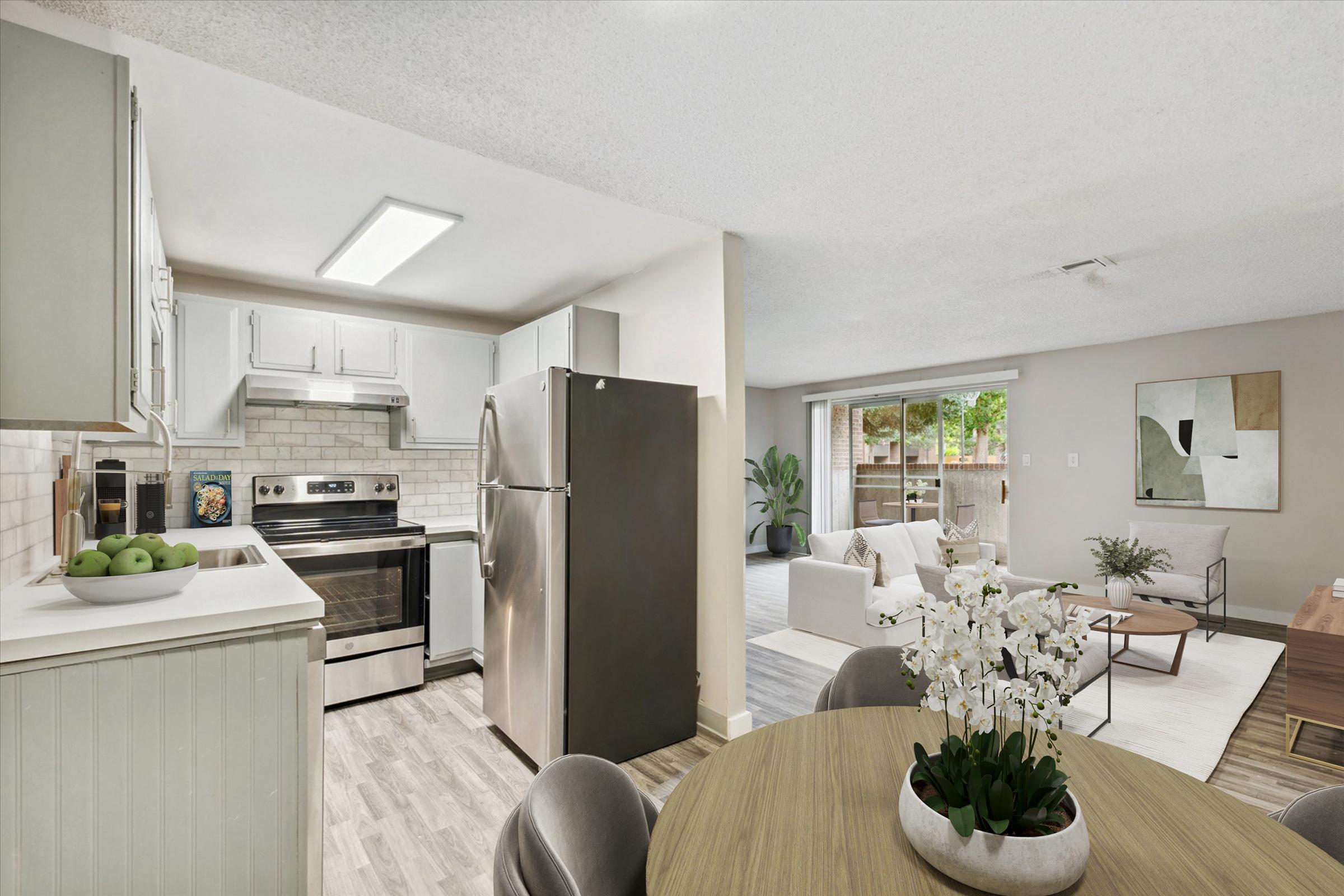 A modern kitchen with stainless steel appliances, including a refrigerator and oven, is adjacent to a cozy living area. The living space features a light-colored sofa, a coffee table, and plants, with large windows letting in natural light. The decor is contemporary and inviting.