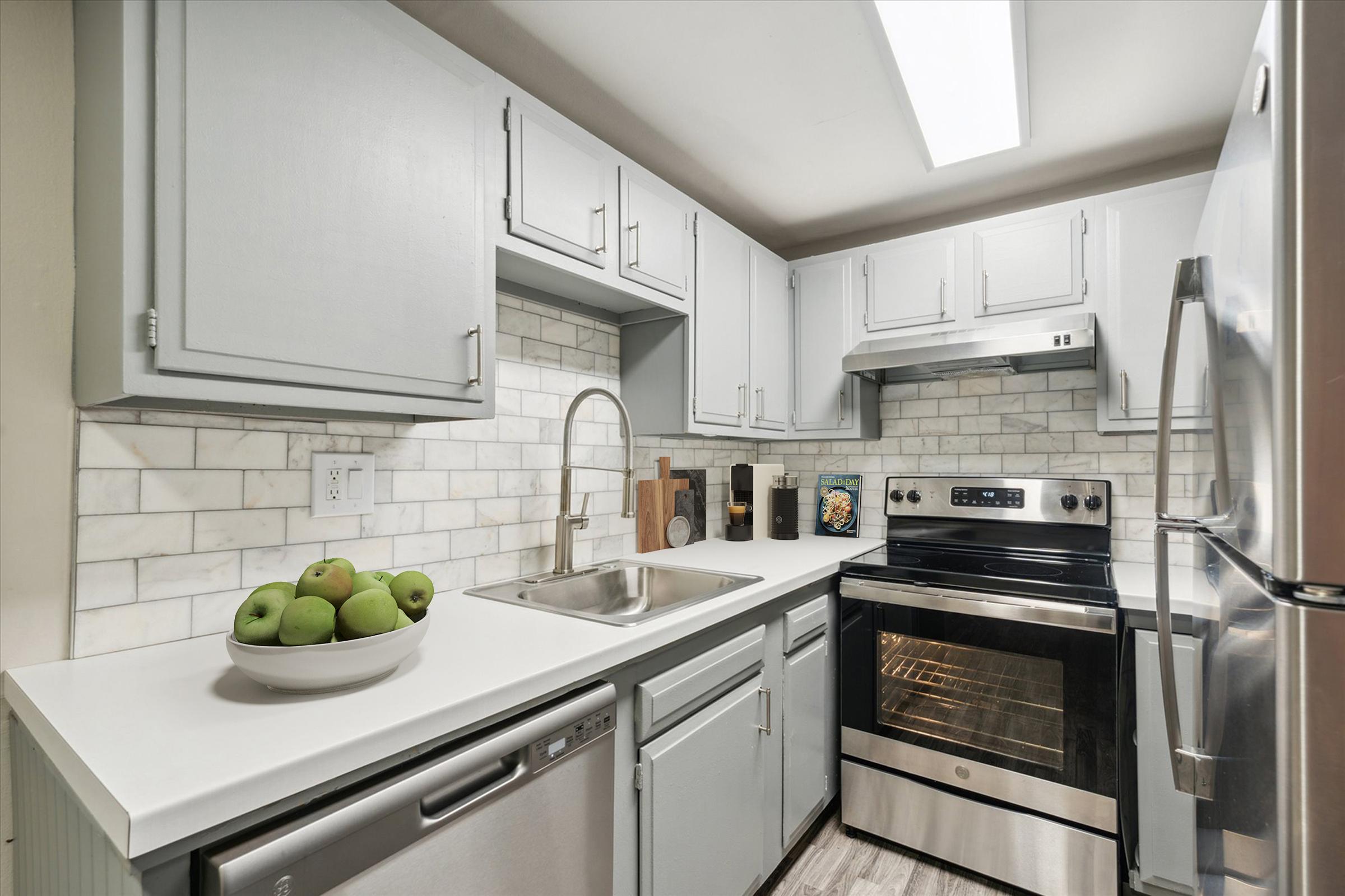 Modern kitchen featuring light gray cabinets, a stainless steel sink, and a flat-top electric stove. A bowl of green apples sits on the countertop, complemented by a tiled backsplash. Stainless steel appliances, including a refrigerator and dishwasher, are included in the design, with natural light illuminating the space.