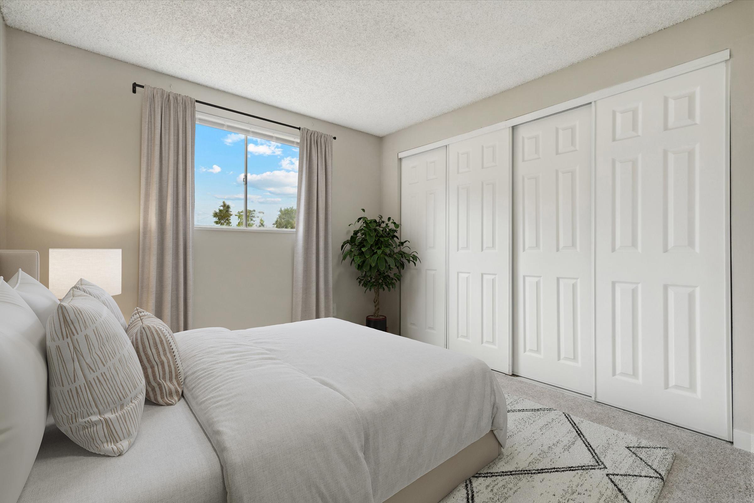 A cozy bedroom featuring a neatly made bed with decorative pillows, a window with sheer curtains allowing natural light, and a potted plant. The walls are a soft beige color, and there are two white closet doors. The room has a modern and inviting atmosphere.
