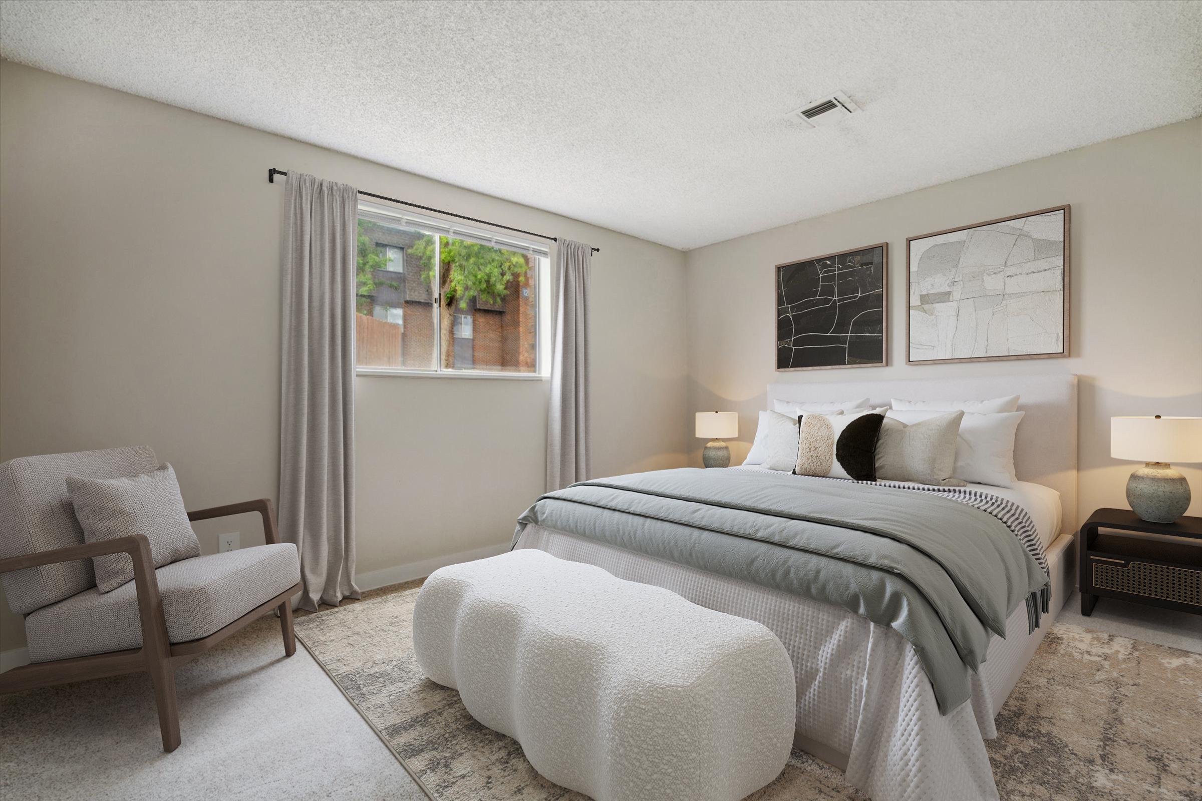 Modern bedroom featuring a queen-sized bed with light blue bedding, a decorative bench at the foot of the bed, and a cozy armchair in the corner. The room is well-lit with natural light coming through a window, and two framed artworks hang above the bed. Neutral-colored walls and a textured carpet enhance the inviting atmosphere.