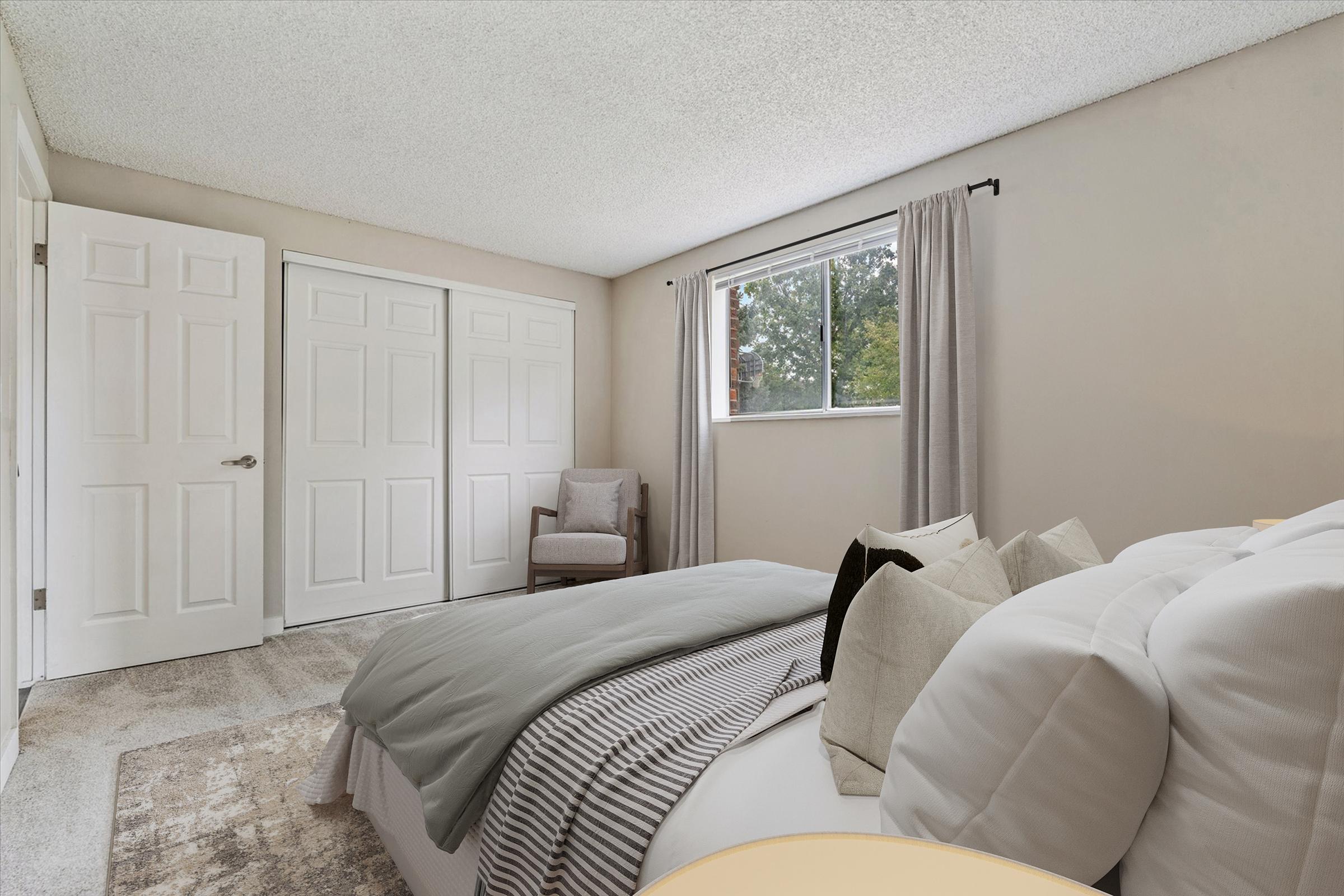 A cozy bedroom featuring a comfortable bed with gray and white bedding, two decorative pillows, and a side table. One corner has a single armchair and a window that provides natural light. The walls are painted light colors, and there are two white closet doors in the background.