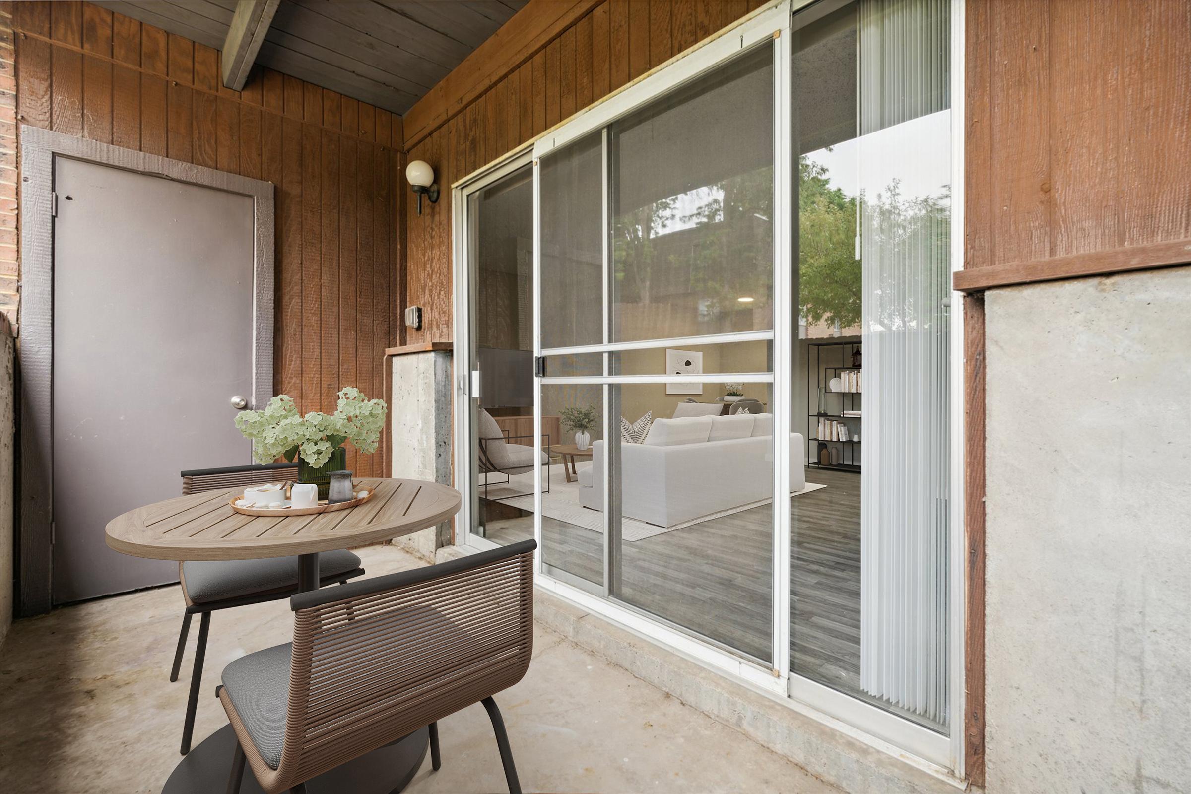 An outdoor patio featuring a round table set with a decorative plant and small plates. A sliding glass door provides a view into a bright living area with a cream-colored sofa and modern decor. The surrounding walls are a mix of wood and concrete, creating a cozy atmosphere.