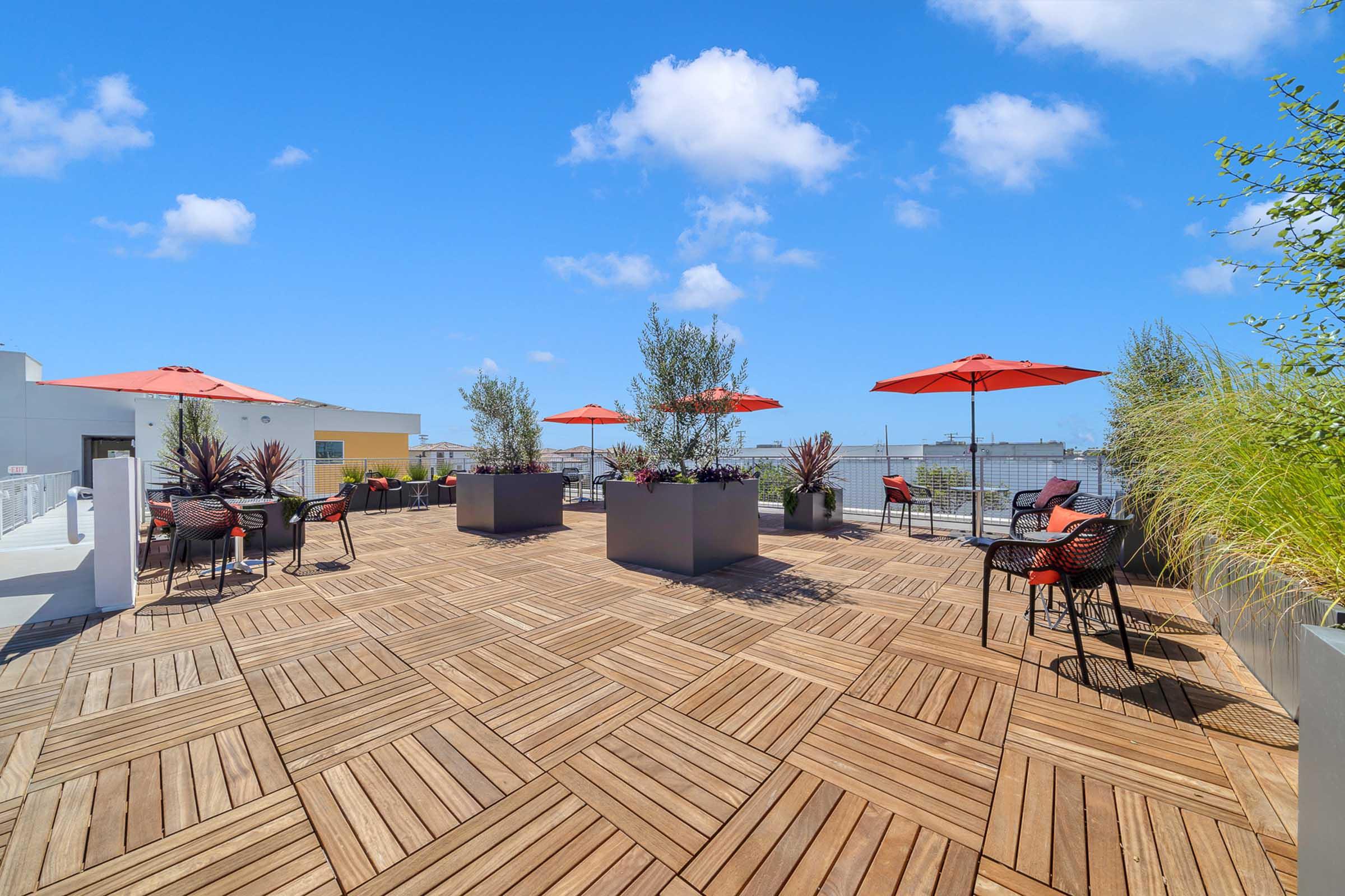 A rooftop terrace featuring wooden flooring, vibrant red umbrellas, and potted plants. The area includes black outdoor chairs around tables, with a clear blue sky in the background and scattered clouds, creating a serene outdoor space for relaxation or gatherings.