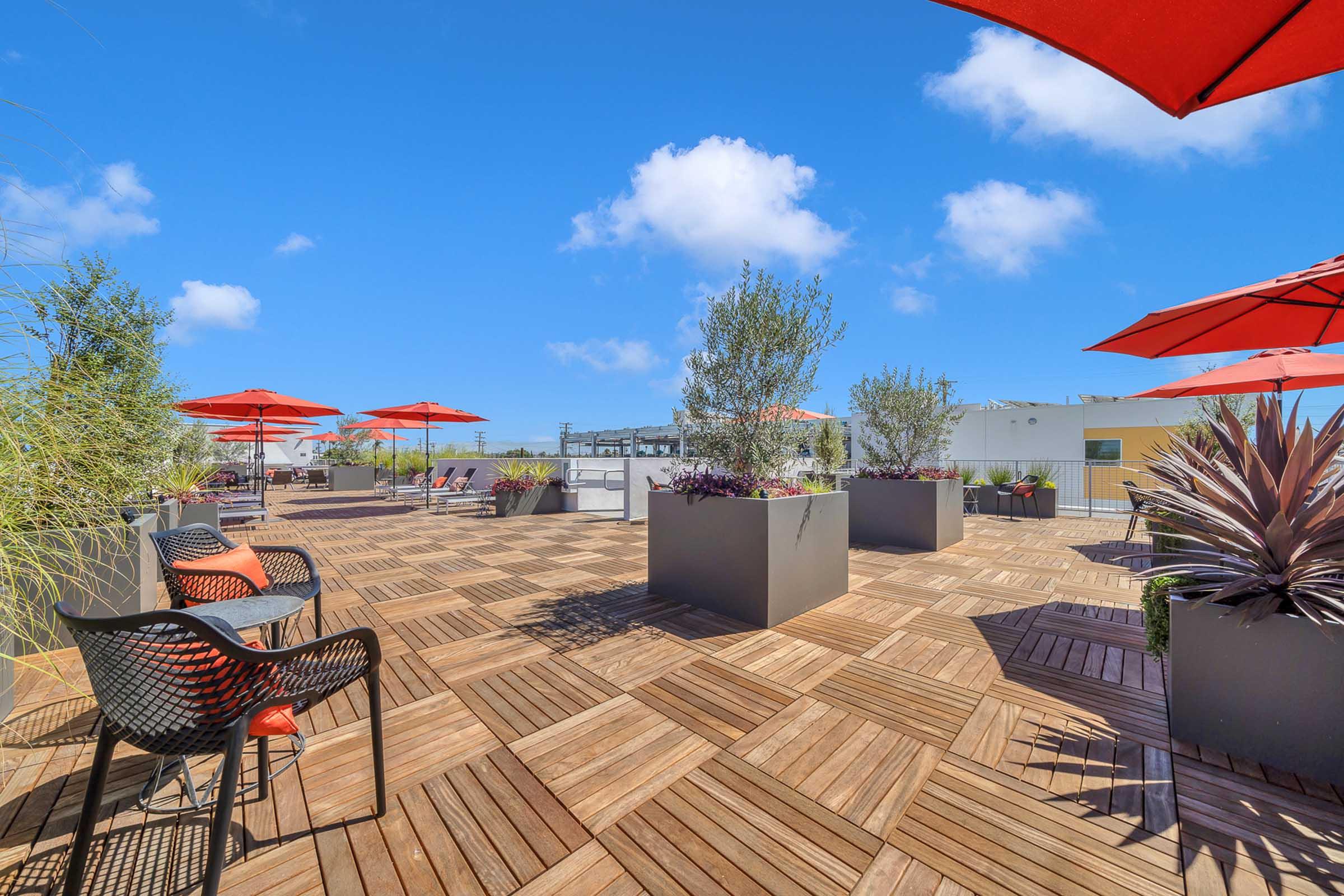 A rooftop terrace featuring wooden decking, large planters with plants, and bright red umbrellas for shade. Black chairs with orange cushions are arranged around the space, under a clear blue sky with a few fluffy clouds. The setting suggests a vibrant outdoor relaxation area.