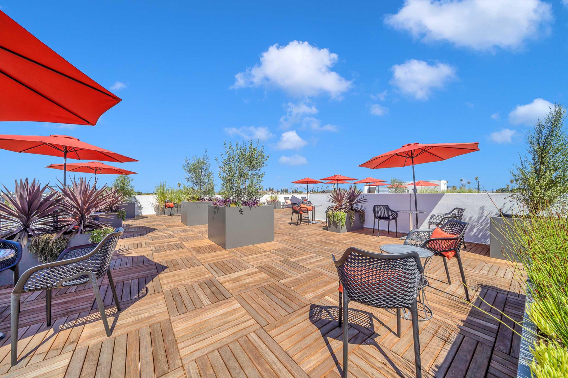 Rooftop terrace featuring wooden flooring, potted plants, and several seating areas with black and gray chairs. Bright red umbrellas provide shade on a sunny day with a clear blue sky and fluffy white clouds in the background.