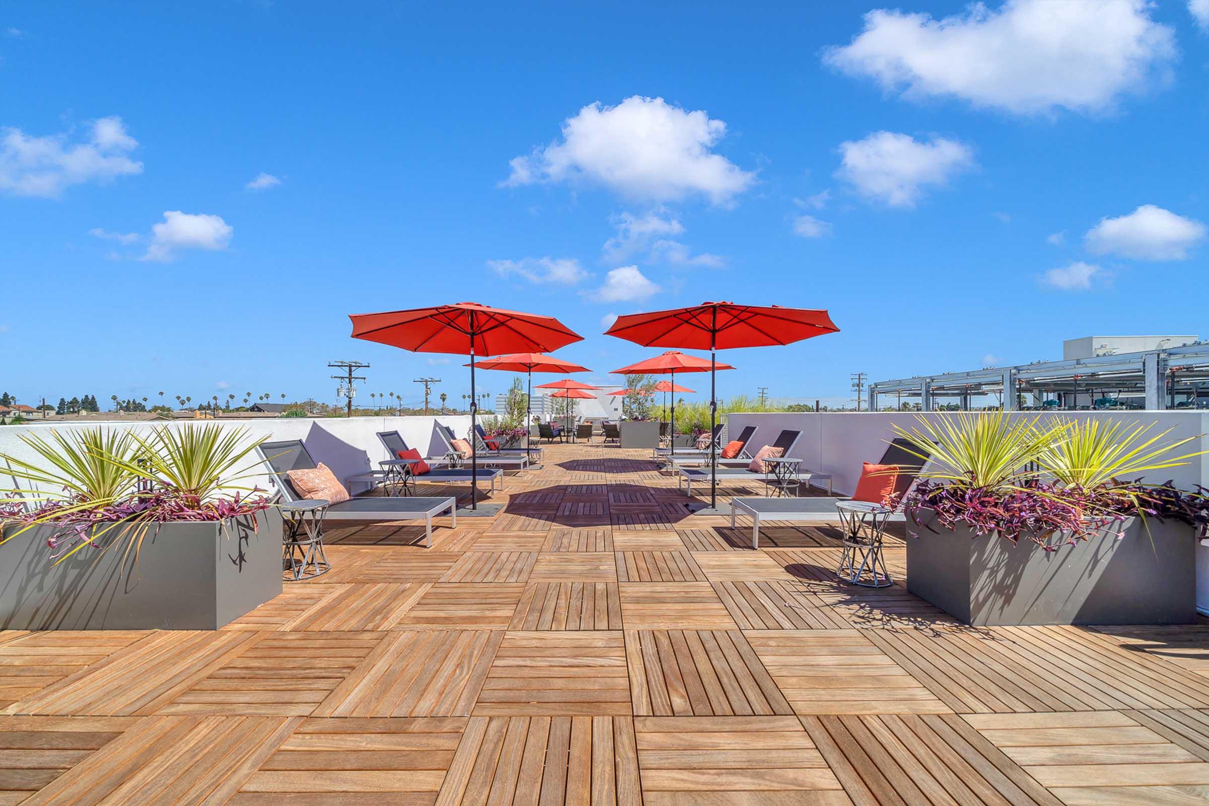 A sunny rooftop terrace featuring wooden deck flooring, with several lounge chairs and orange umbrellas providing shade. Colorful plants in modern planters add greenery, and the sky is bright blue with fluffy clouds. The overall atmosphere is relaxed and inviting.