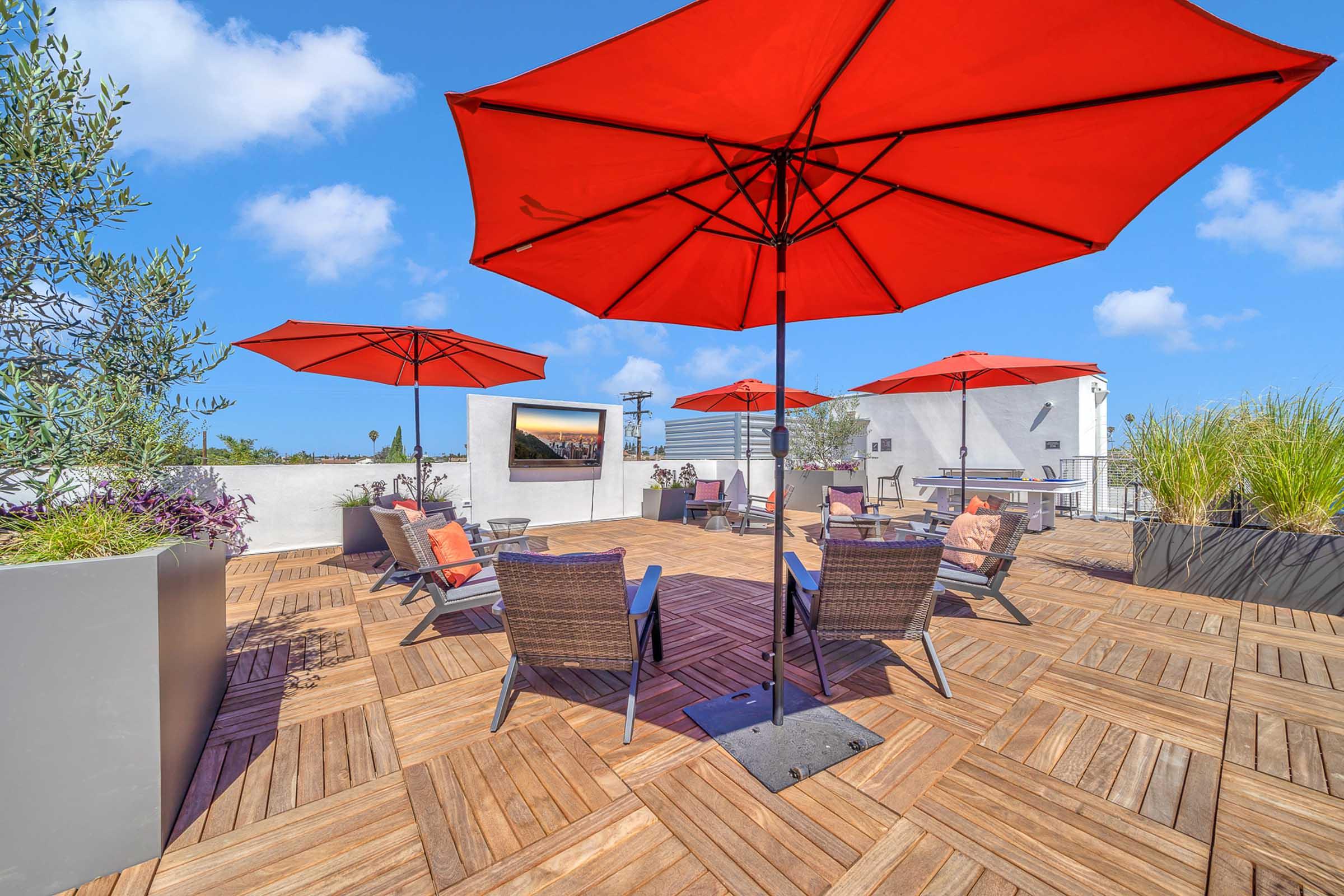 A rooftop patio featuring wooden decking, several red umbrellas providing shade, and lounge chairs arranged around small tables. Green plants and a television screen are visible in the background under a clear blue sky.