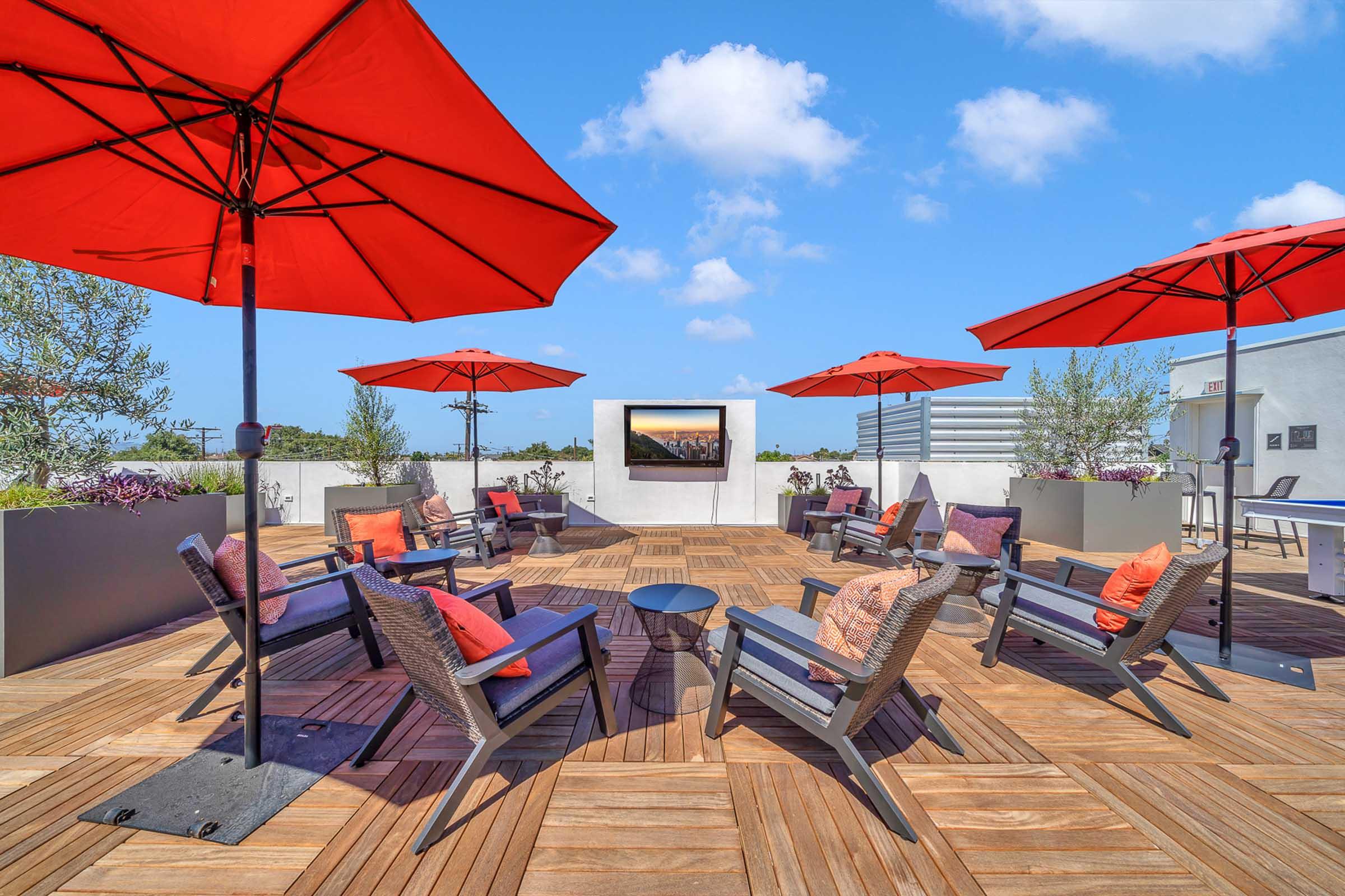 A rooftop terrace featuring several stylish lounge chairs and small tables, adorned with orange and red cushions. Bright red umbrellas provide shade, and a modern screen is mounted on the wall. The backdrop includes blue skies and a few scattered clouds, creating a vibrant outdoor relaxation space.