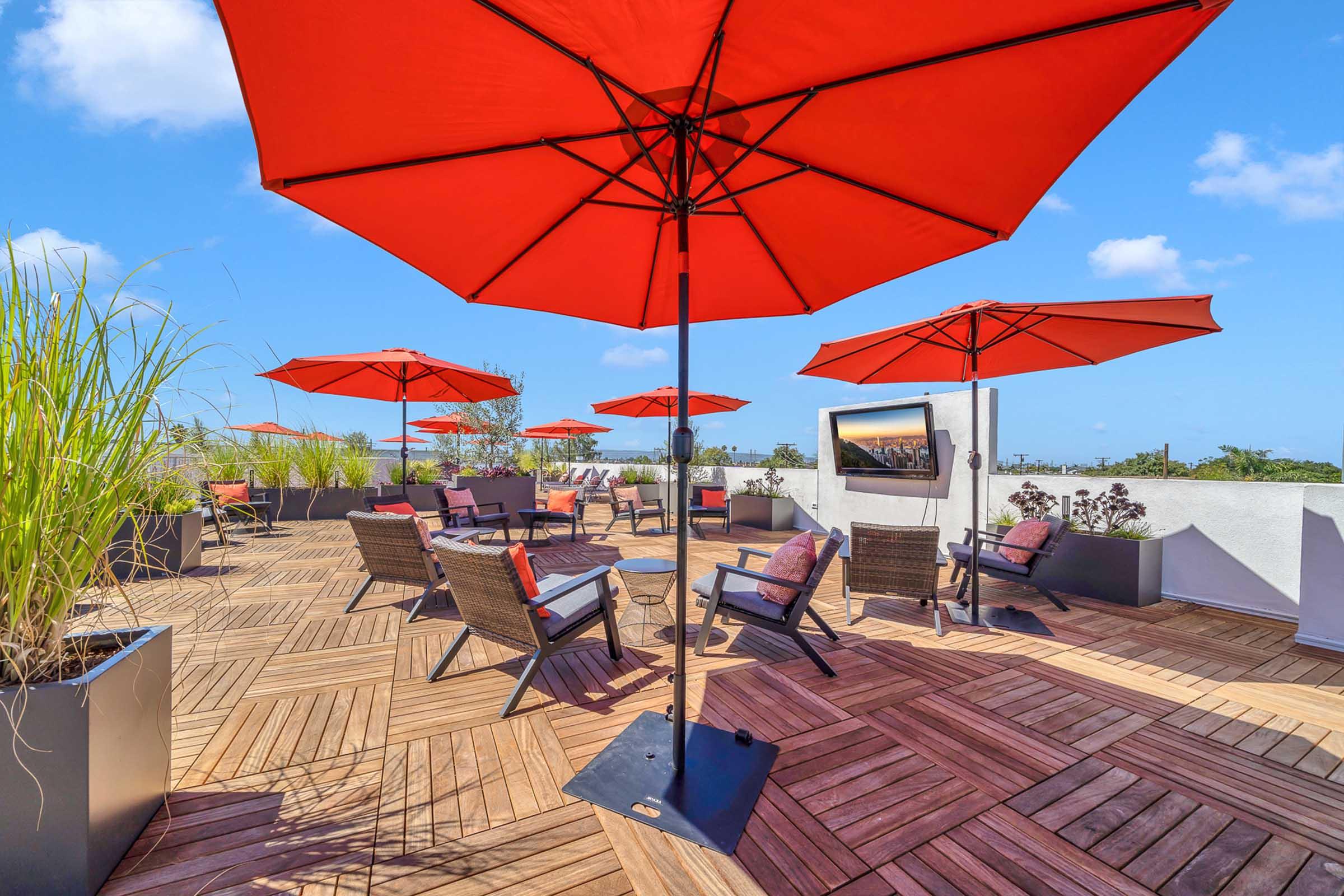 A rooftop terrace featuring wooden flooring and several seating areas, each with wicker chairs and red umbrellas providing shade. Lush green plants in stylish pots add to the ambiance, while a bright blue sky complements the sunny atmosphere.
