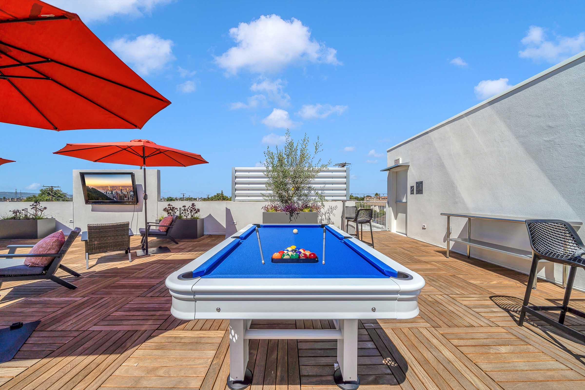 A rooftop lounge area featuring a blue pool table with a full set of pool balls arranged, surrounded by stylish patio furniture and red umbrellas. A planter with greenery is visible in the background, along with a view of the sky and distant buildings.