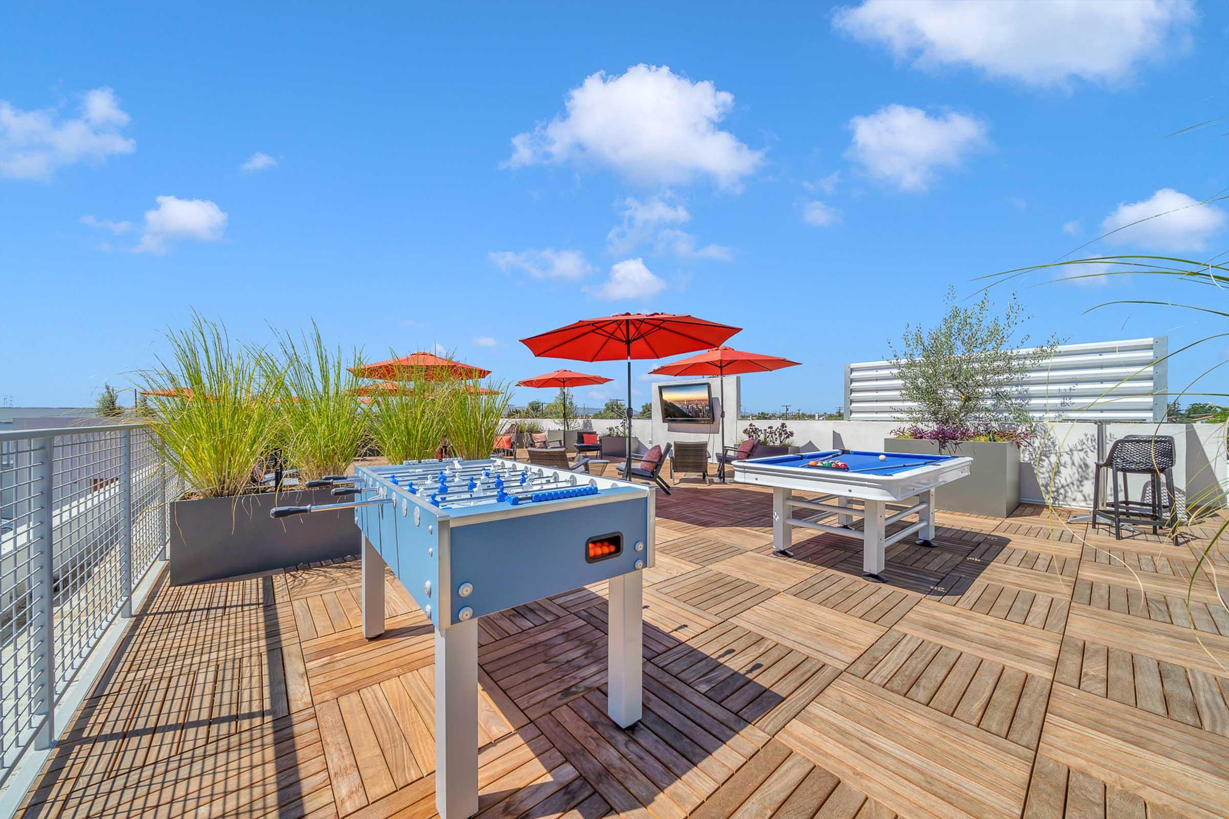 Rooftop terrace with wooden flooring, featuring a foosball table and a pool table. Brightly colored red umbrellas provide shade, surrounded by ornamental grasses and small plants. Clear blue sky with a few fluffy clouds creates a vibrant outdoor space for relaxation and recreation.