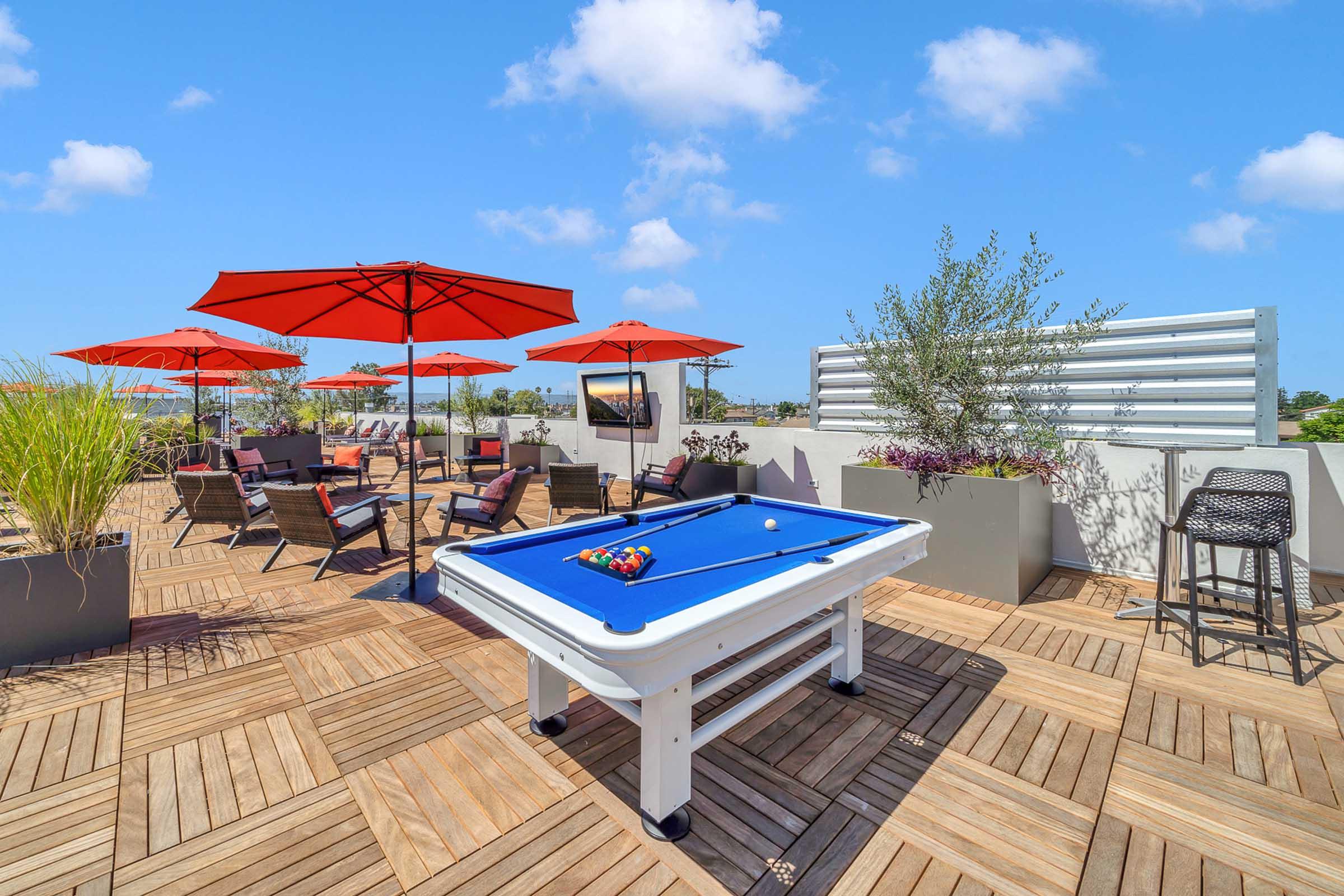 A rooftop terrace featuring a blue pool table, surrounded by red umbrellas and lounge chairs. There are planters with greenery, and a scenic view under a bright blue sky with fluffy clouds. The space is designed for relaxation and entertainment.