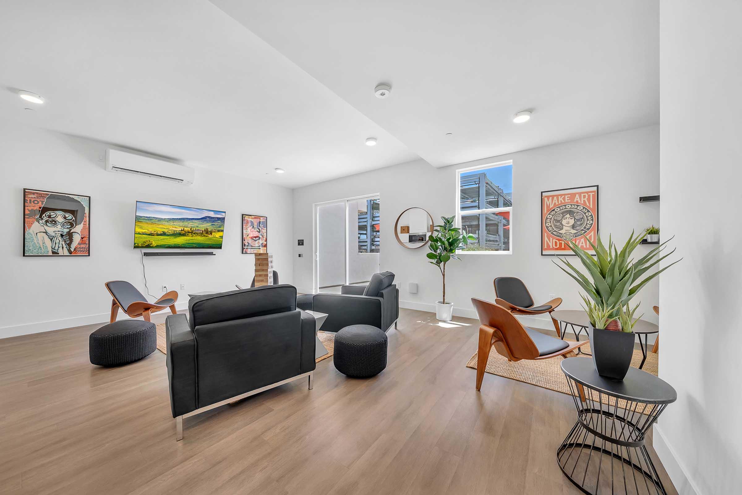 A modern living room featuring a large flat-screen TV on the wall, stylish seating including black sofas and wooden chairs, a decorative plant, and artistic wall posters. Natural light streams in through large windows, creating a bright and inviting atmosphere. A coffee table and area rug complete the cozy setting.