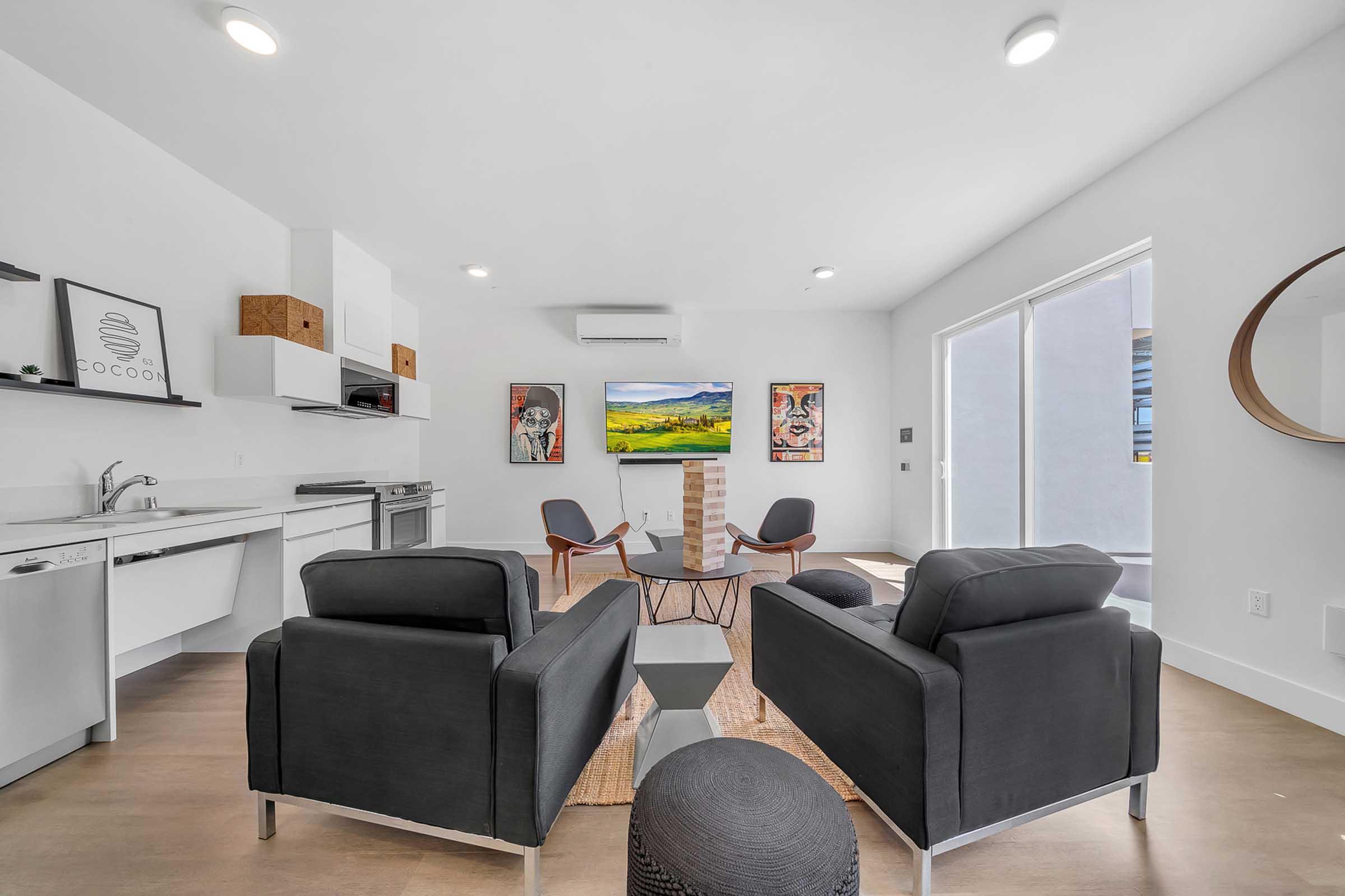 A modern living room featuring two black armchairs and a small black table. There is a television mounted on the wall displaying a landscape image, surrounded by decorative artwork. The kitchen area is visible with minimalist cabinetry and appliances, and large windows allow natural light to fill the space.