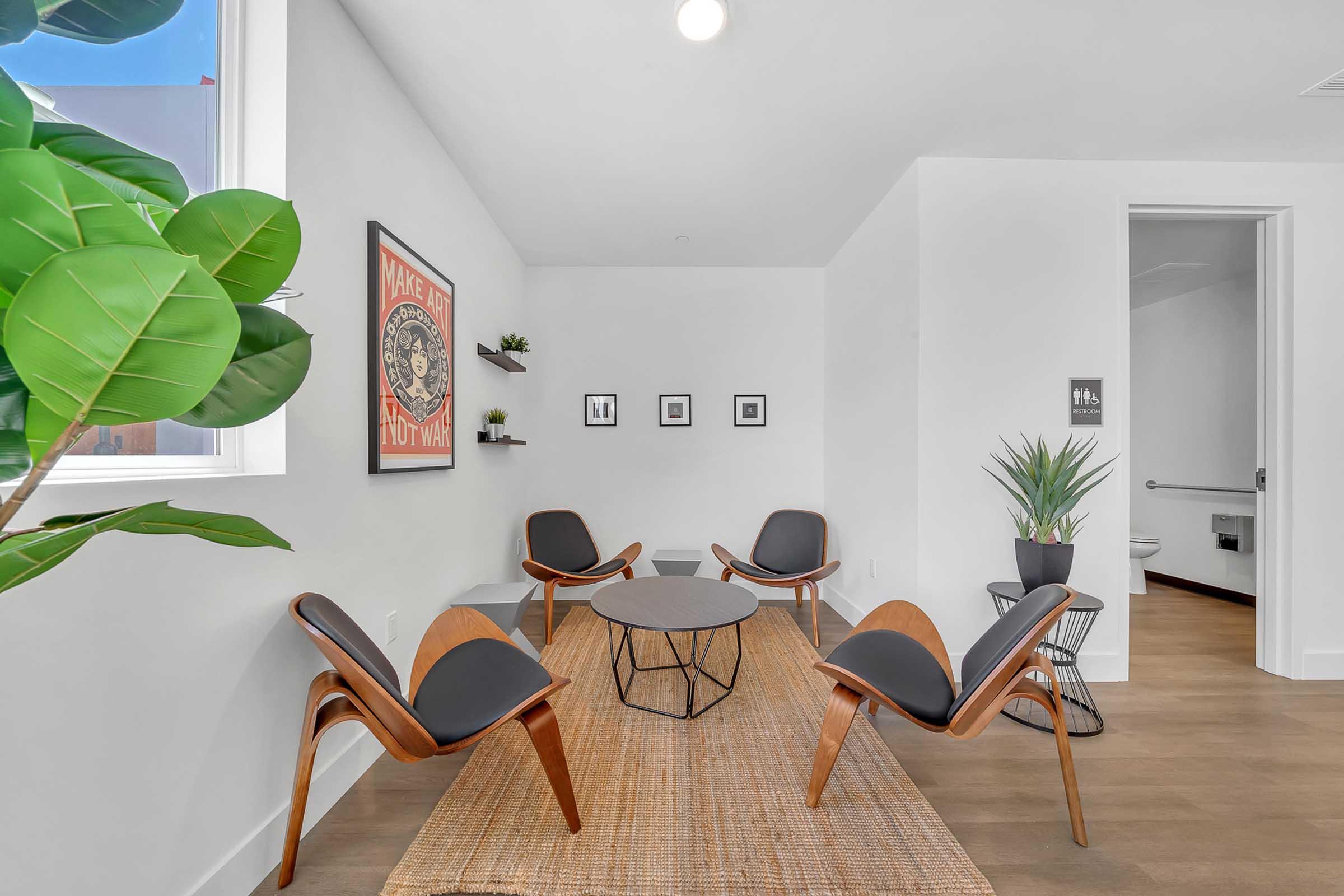 A modern, minimalist waiting room featuring four wooden chairs surrounding a circular black table on a woven rug. Two small wall shelves hold decorative items, and framed pictures are displayed on the wall. A large window lets in natural light, and a green plant adds a touch of nature to the space.