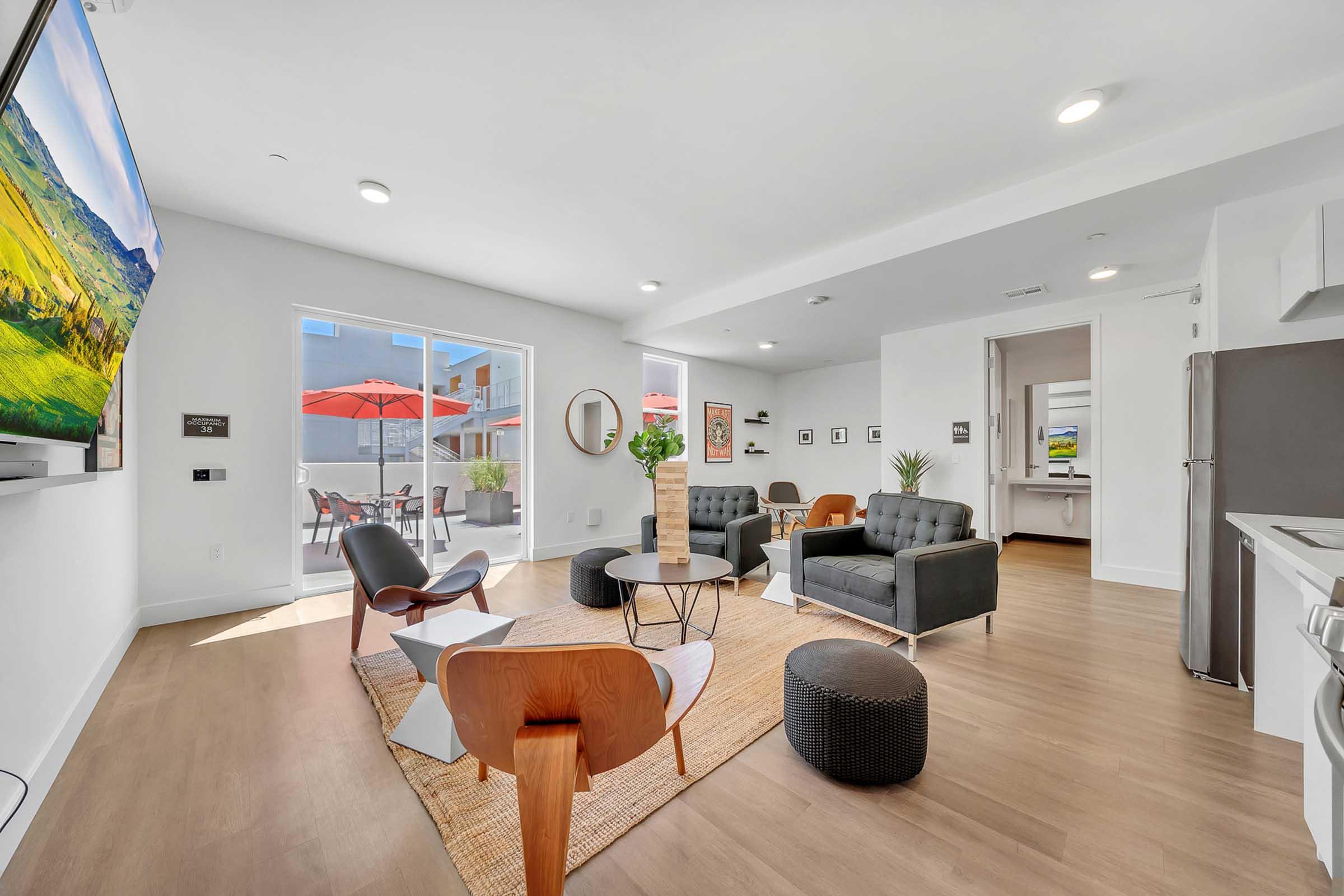 A modern living space featuring a bright, open layout. Furnished with stylish black and wood chairs, a round coffee table, and a decorative rug. Large windows allow natural light, with a view of an outdoor area with red umbrellas. A sleek TV and contemporary décor complete the warm ambiance.