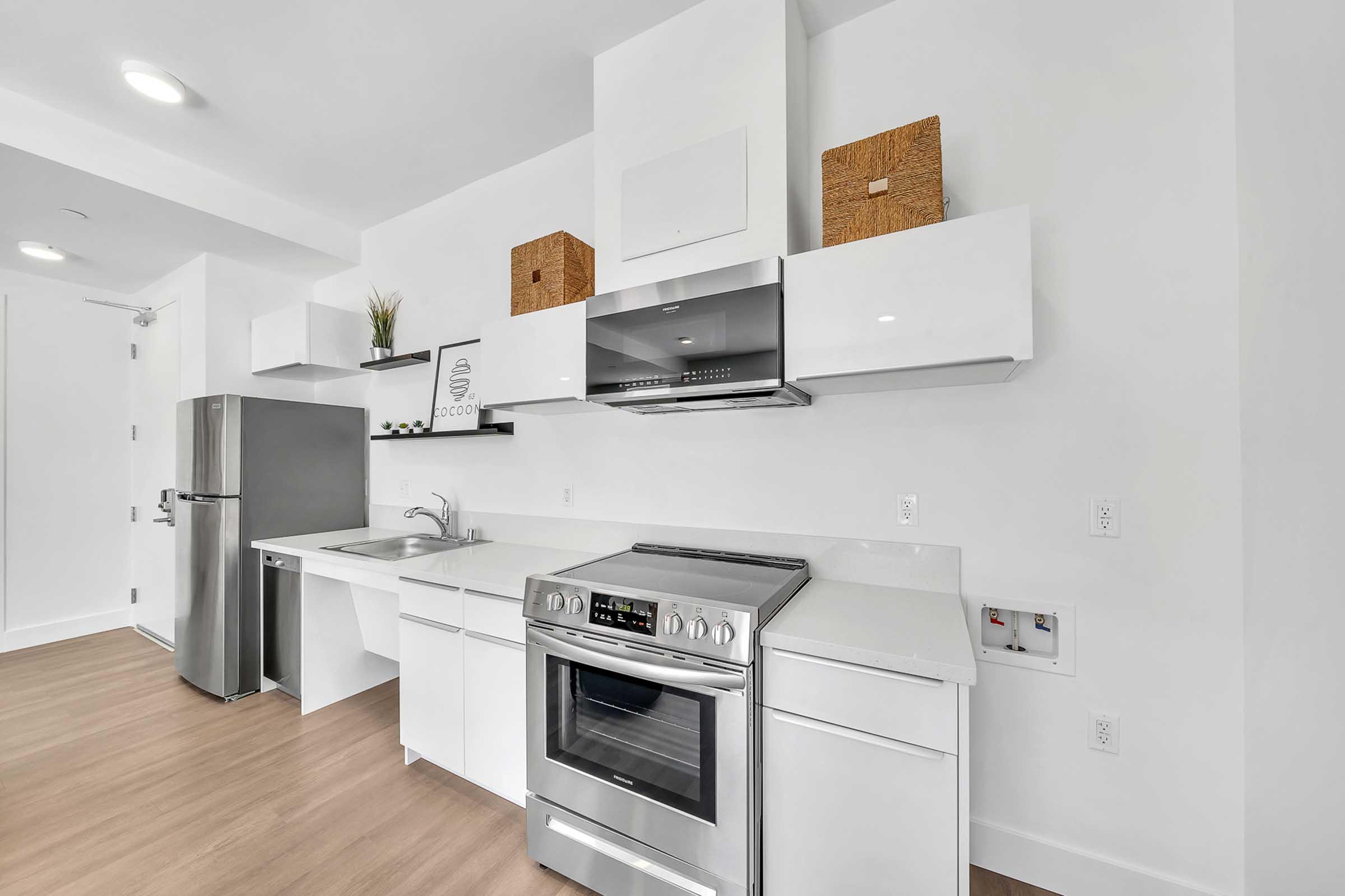 Modern kitchen featuring sleek white cabinetry, stainless steel appliances including a refrigerator and oven, a sink with a minimalist faucet, and light wood flooring. Decorative boxes and a plant add a touch of style to the countertop, and the space is well-lit with recessed lighting.