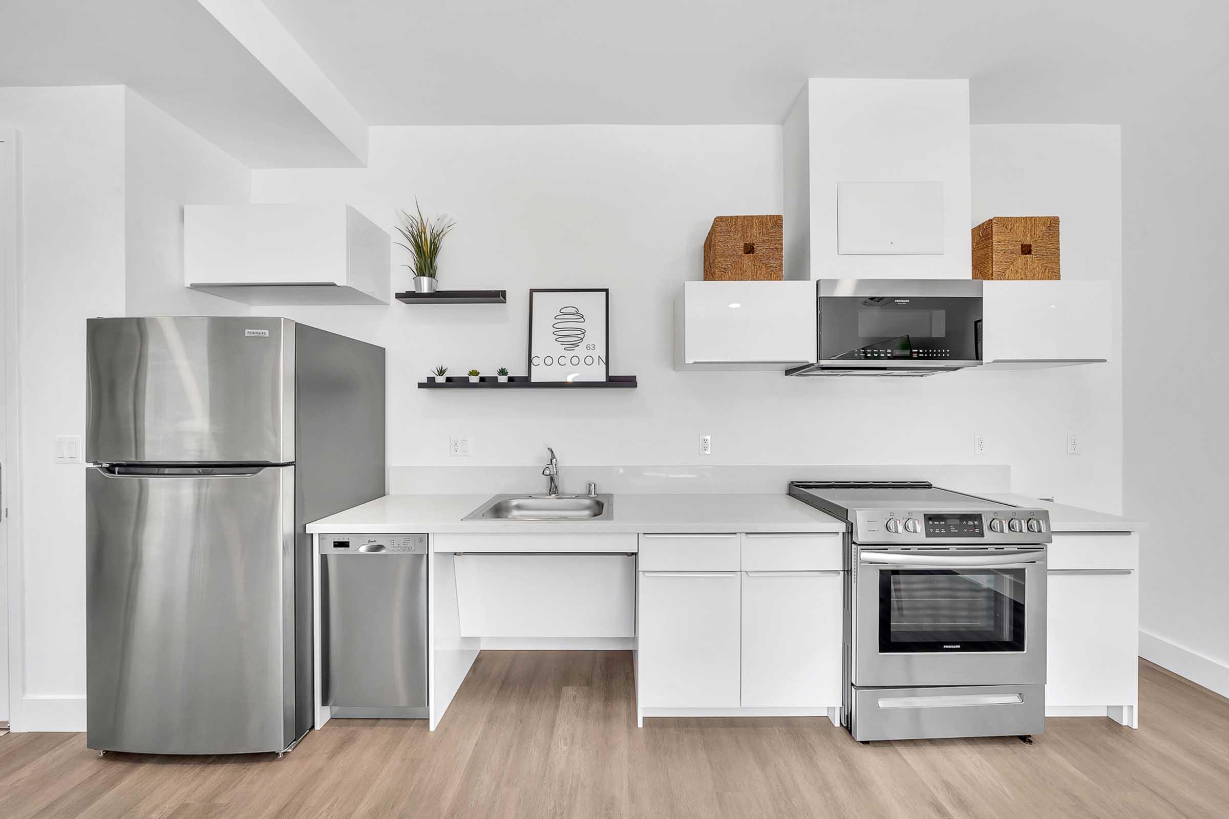 Modern kitchen featuring stainless steel appliances, including a refrigerator, dishwasher, and oven. The sleek white cabinetry is complemented by open shelves displaying decorative items. The space has a minimalist design with light wood flooring and a neutral color palette. A framed print is visible on the wall.
