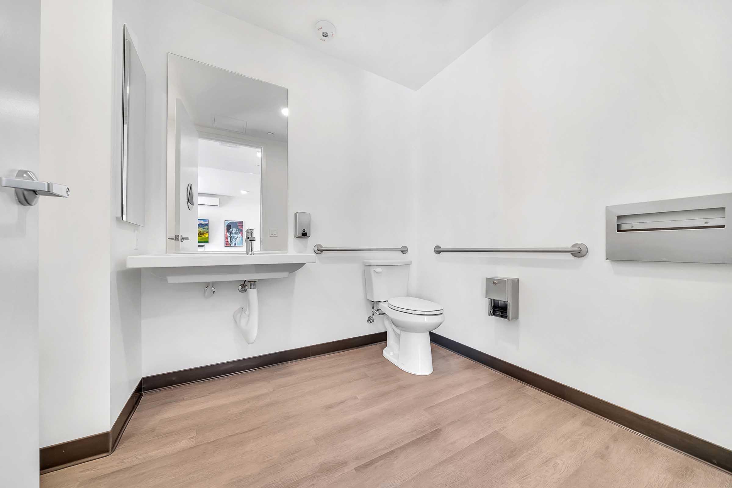 A clean, minimal restroom featuring light-colored walls, a mirror above a sink, toilet, and grab bars. The floor is wood-like, and there’s a mail slot on the wall. Soft overhead lighting enhances the bright atmosphere.