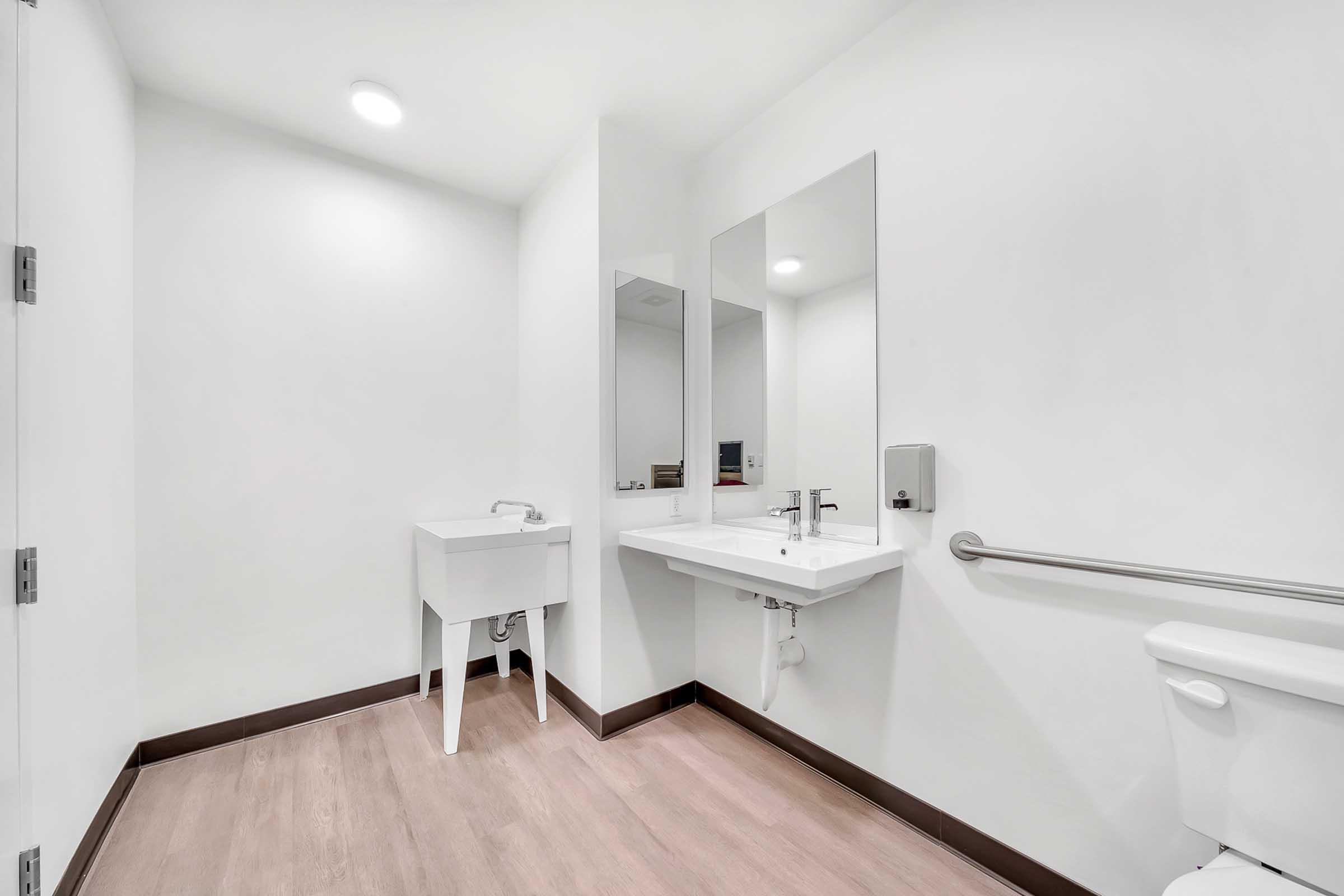 A clean, modern restroom featuring a white sink with a mirror above, a wall-mounted soap dispenser, and a toilet. The walls are painted white, and the floor is light wood. There's a grab bar for accessibility, contributing to the spacious, bright design.